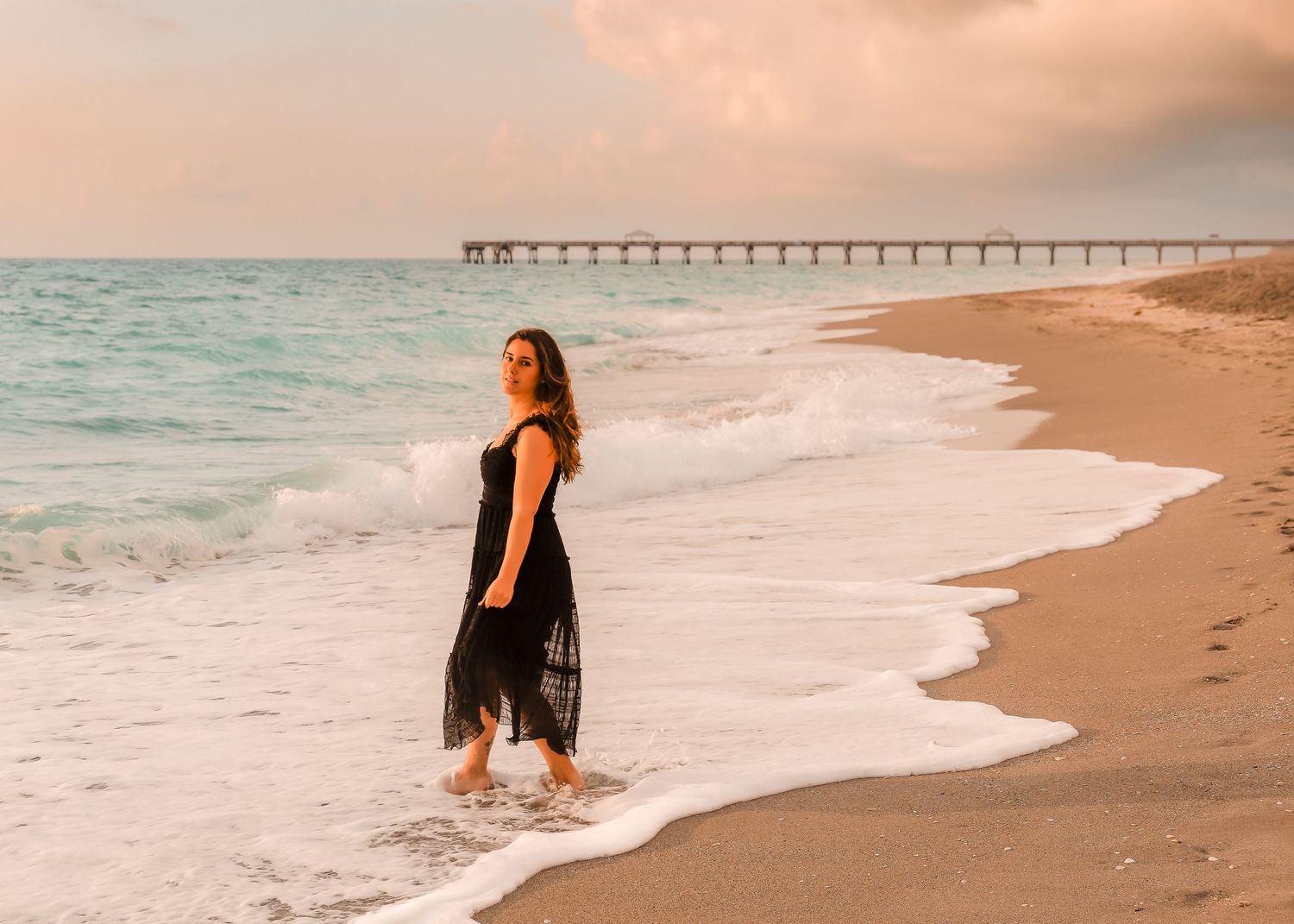 Woman in black dress walking on sandy beach at sunset with ocean waves and pier in background.