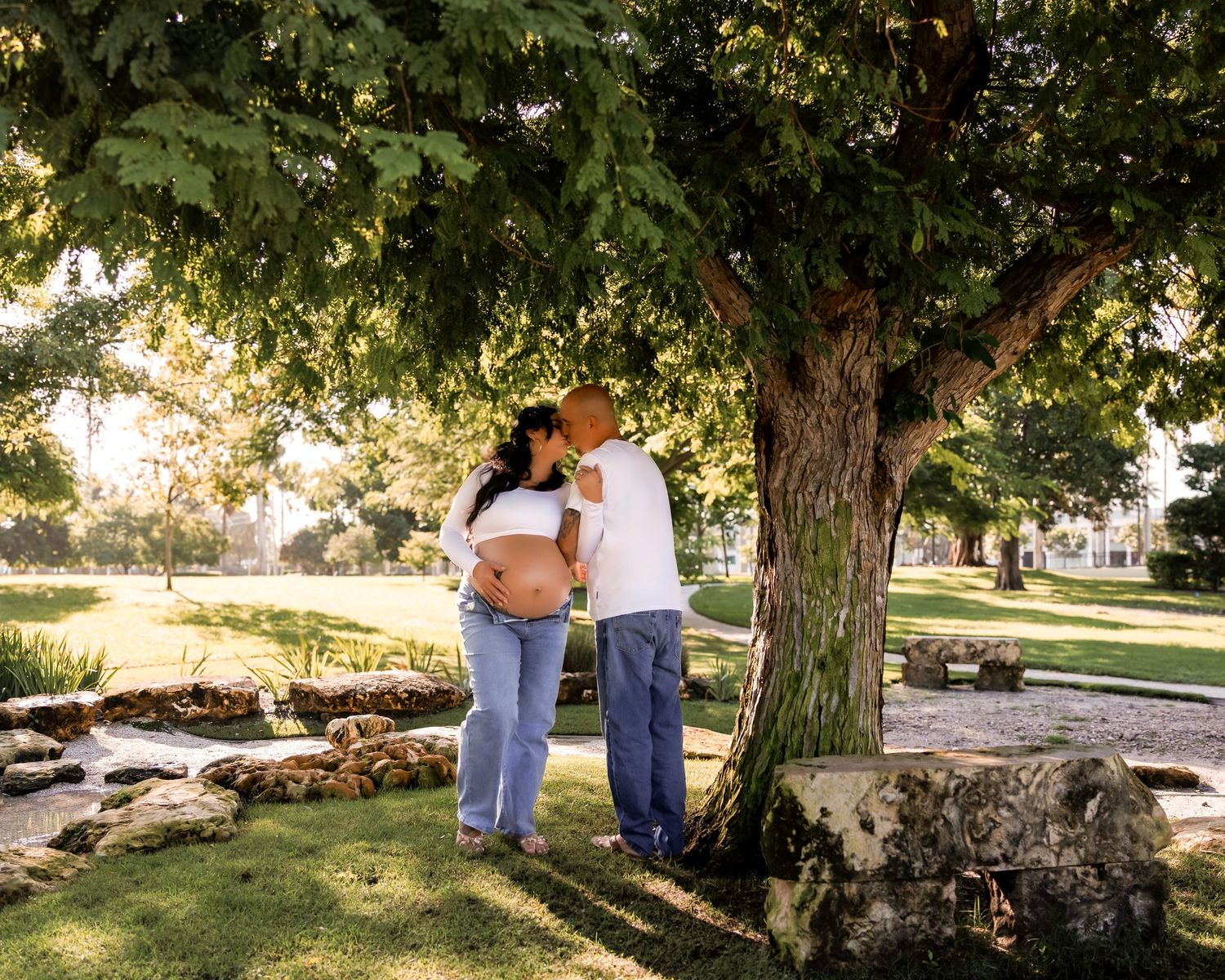 Pregnant couple sharing a tender moment under a large tree in a sunny park, maternity photo session outdoors.