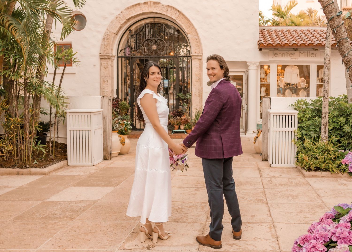 Couple holding hands in front of an elegant Mediterranean-style venue, woman in white dress, man in burgundy blazer.