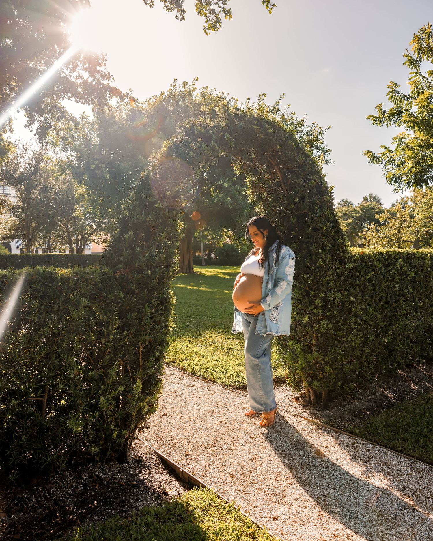 Pregnant woman standing under a garden arch in golden sunlight, wearing jeans and a denim jacket.