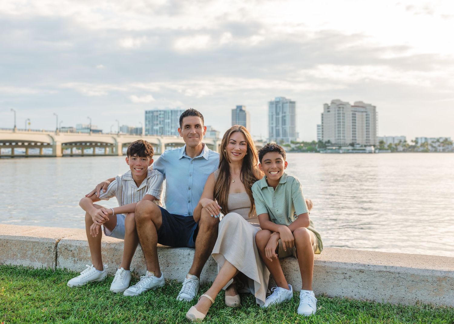Five casual friends lounge together on grass enjoying ocean views and city skyline at sunset.