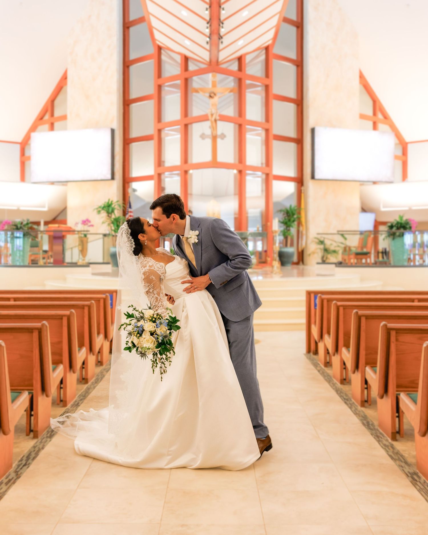 Bride and groom share first kiss in elegant chapel aisle, bride holding white bouquet in stunning wedding gown.
