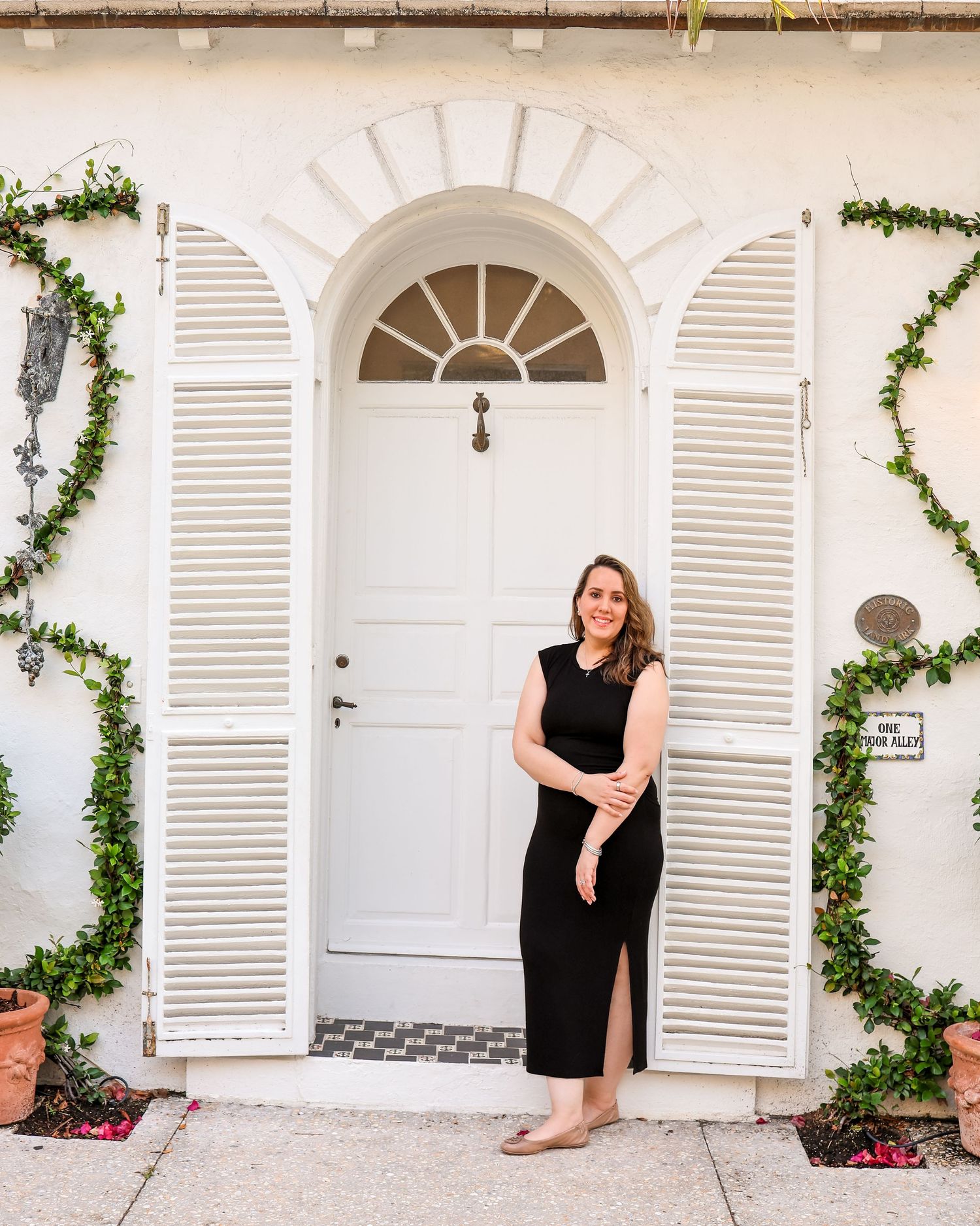 Woman in black dress posing before elegant white arched door with shutters and ivy-covered walls.