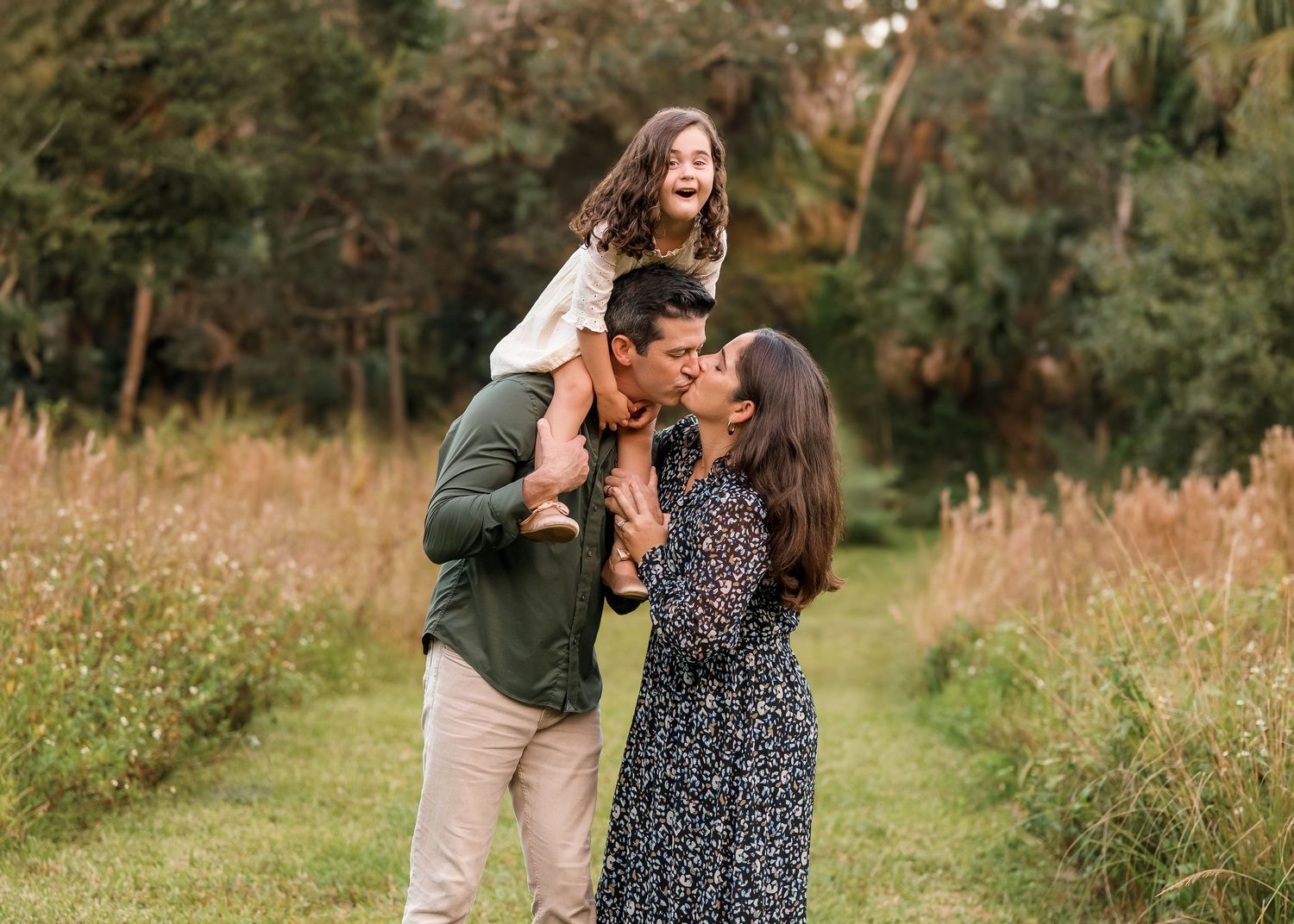 A joyful family of three poses outdoors, parents kissing while daughter sits on dad's shoulders laughing in a scenic field.
