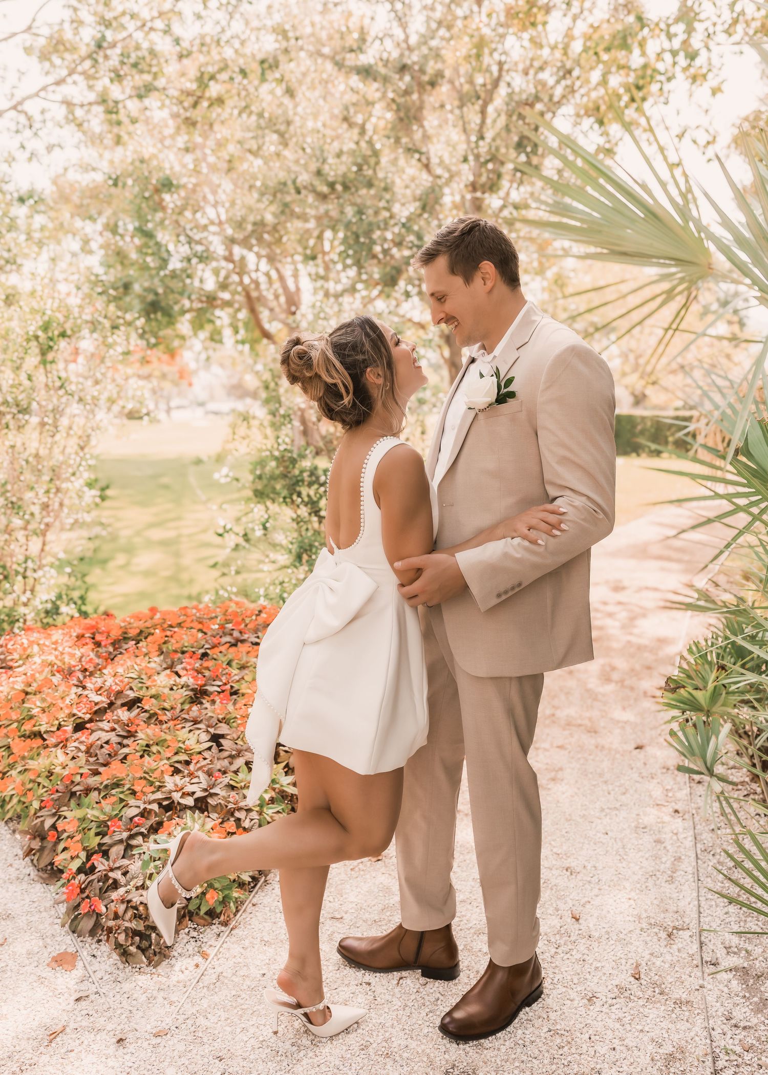 Bride in white mini dress with bow and groom in tan suit share a romantic moment on a garden path.