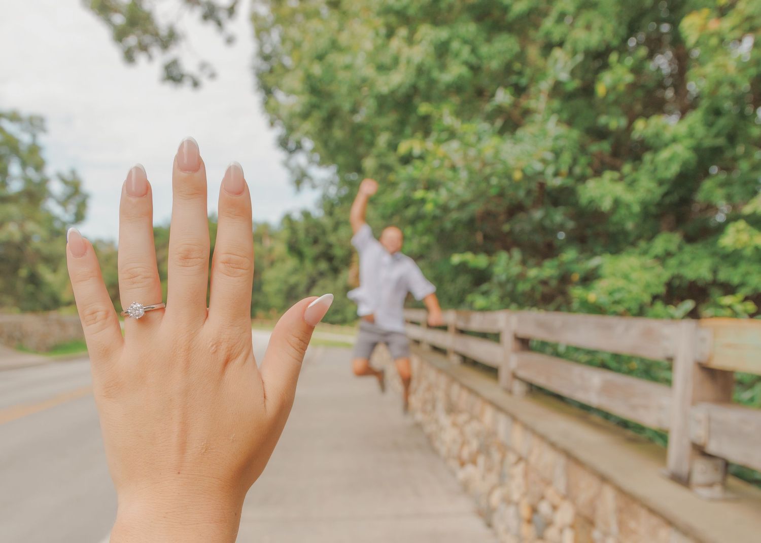 Hand wearing a gold ring held up against a blurry bridge railing on a sunny day.