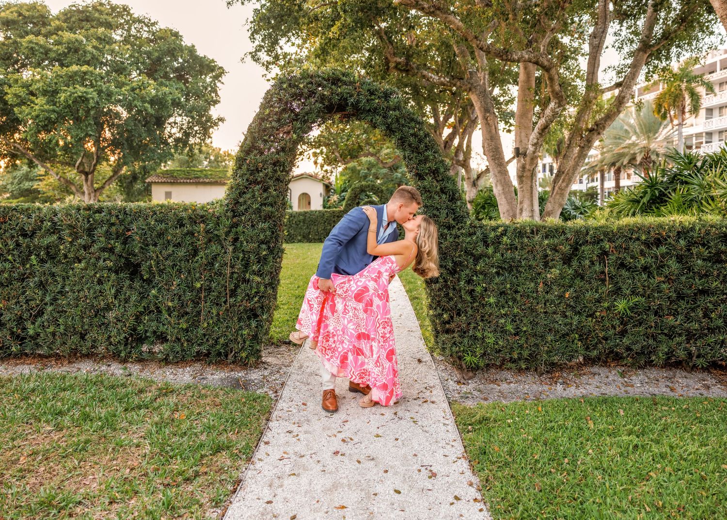 Couple sharing a romantic dip kiss under a lush green hedge archway in a manicured garden at golden hour.