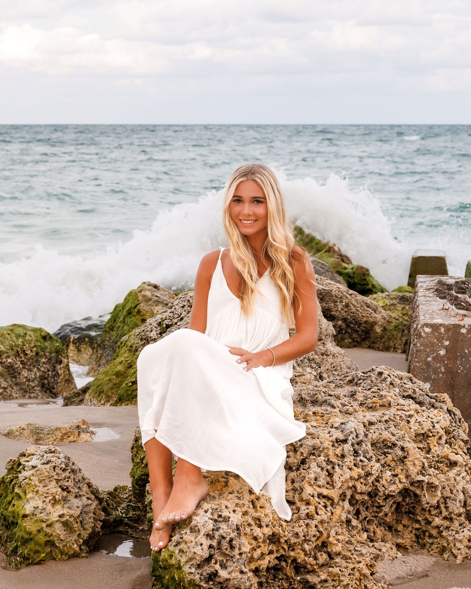 Blonde woman in white maxi dress sitting on mossy rocks by ocean waves at beach.