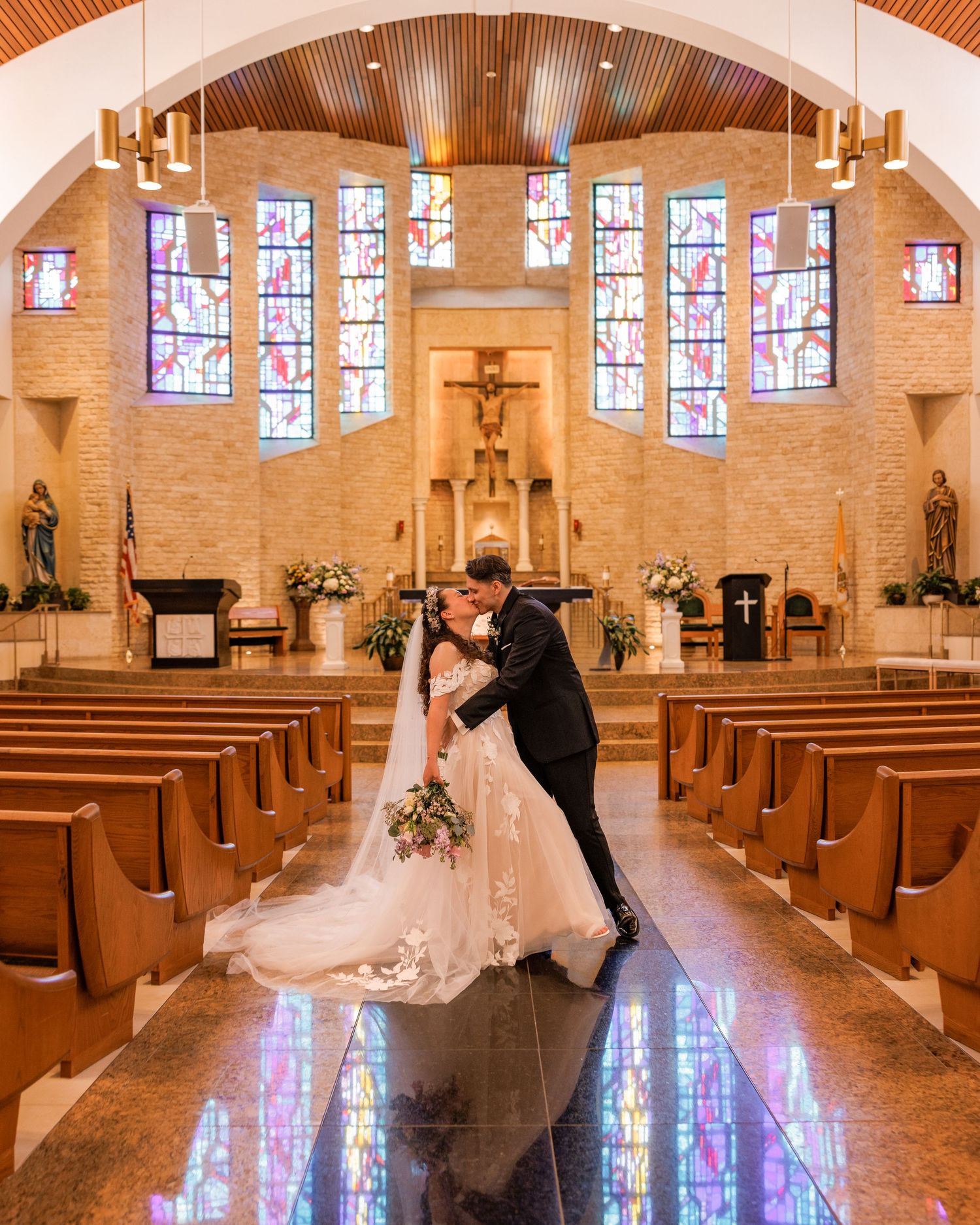 Bride and groom share a romantic kiss at the altar of a stunning Catholic church with stained glass windows.