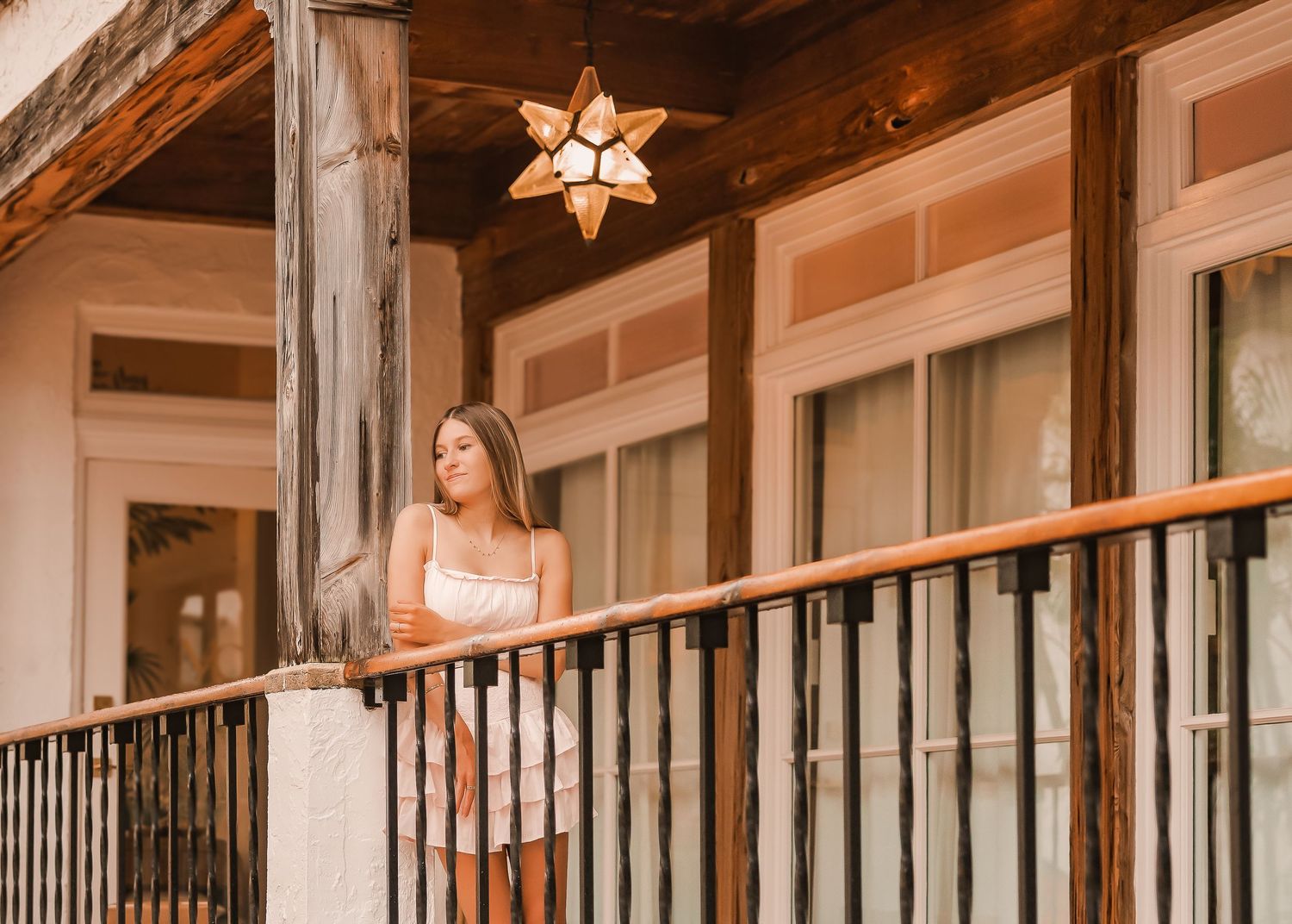 Young woman in white dress standing on a rustic wooden balcony with iron railing and star pendant light.