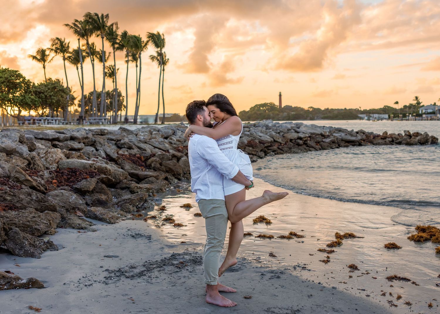 Couple embracing on a sandy beach at sunset with palm trees and rocky shoreline in a tropical setting.