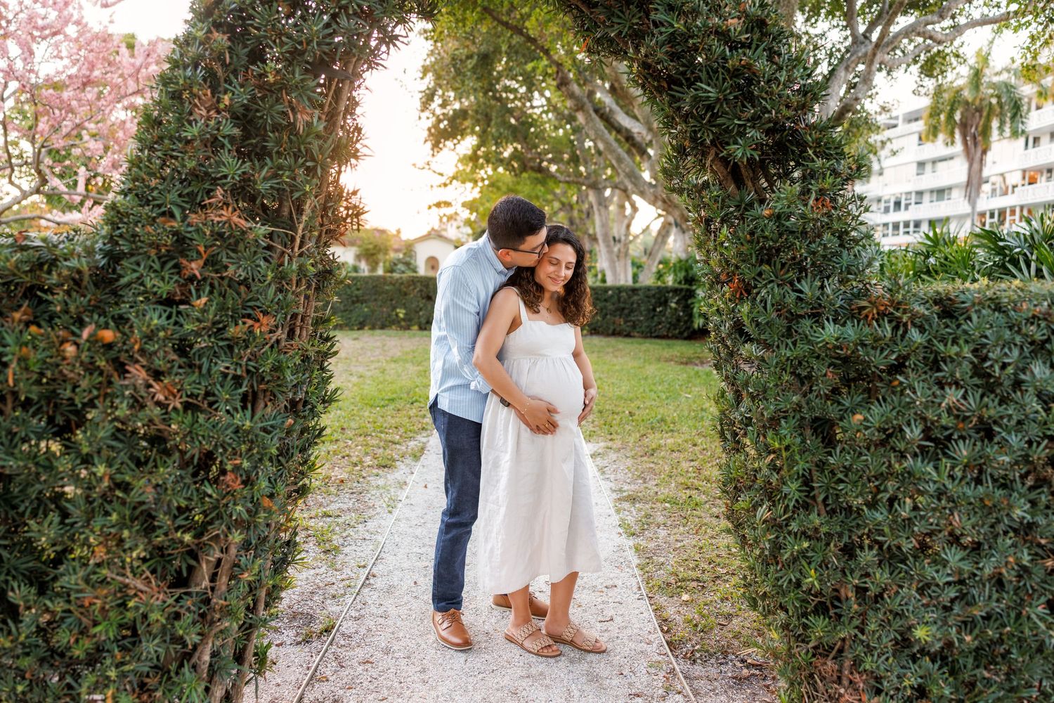 Couple sharing a tender maternity moment under a lush garden archway, man kissing pregnant woman's head outdoors.