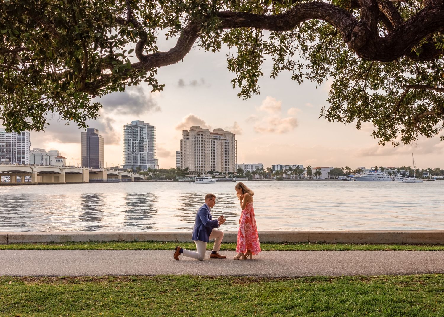 Man proposes on one knee to woman in floral dress by waterfront with city skyline at sunset.
