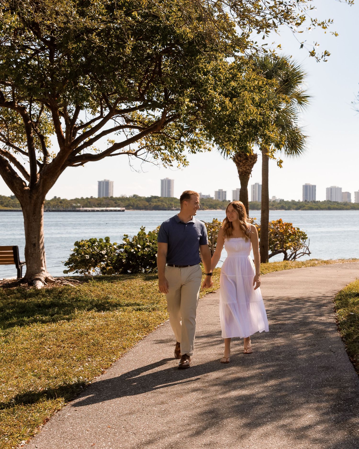 Couple holding hands walking on a sunny waterfront park path with palm trees and city skyline in background.