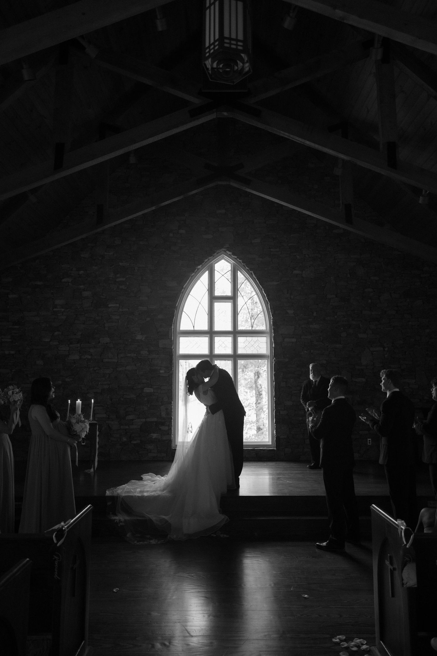 Black and white photo of bride and groom kissing at altar in chapel with gothic arched window.