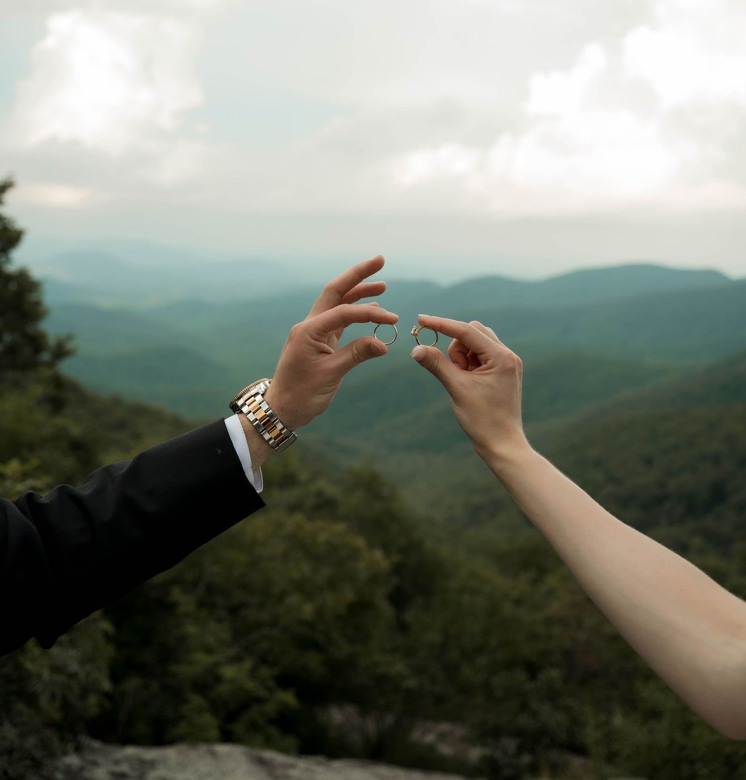 Bride and groom touching fingertips holding wedding ring with mountain landscape backdrop.