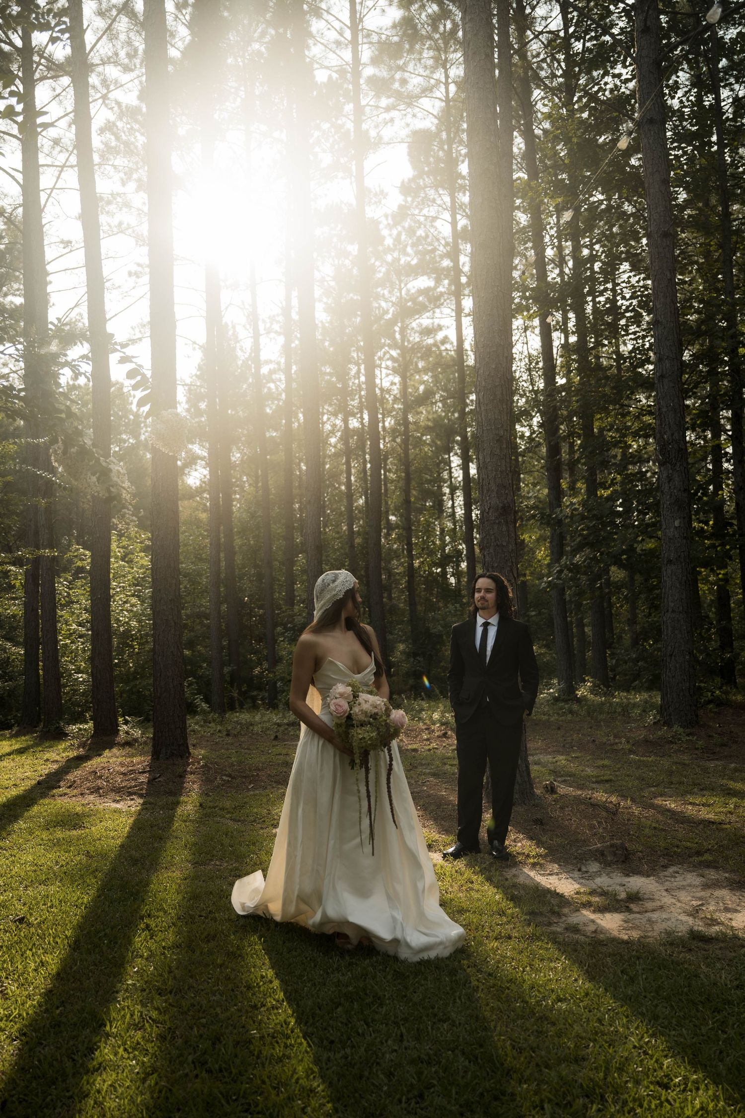 Bride in white dress holding bouquet faces groom in dark suit during golden hour first look in a pine forest.
