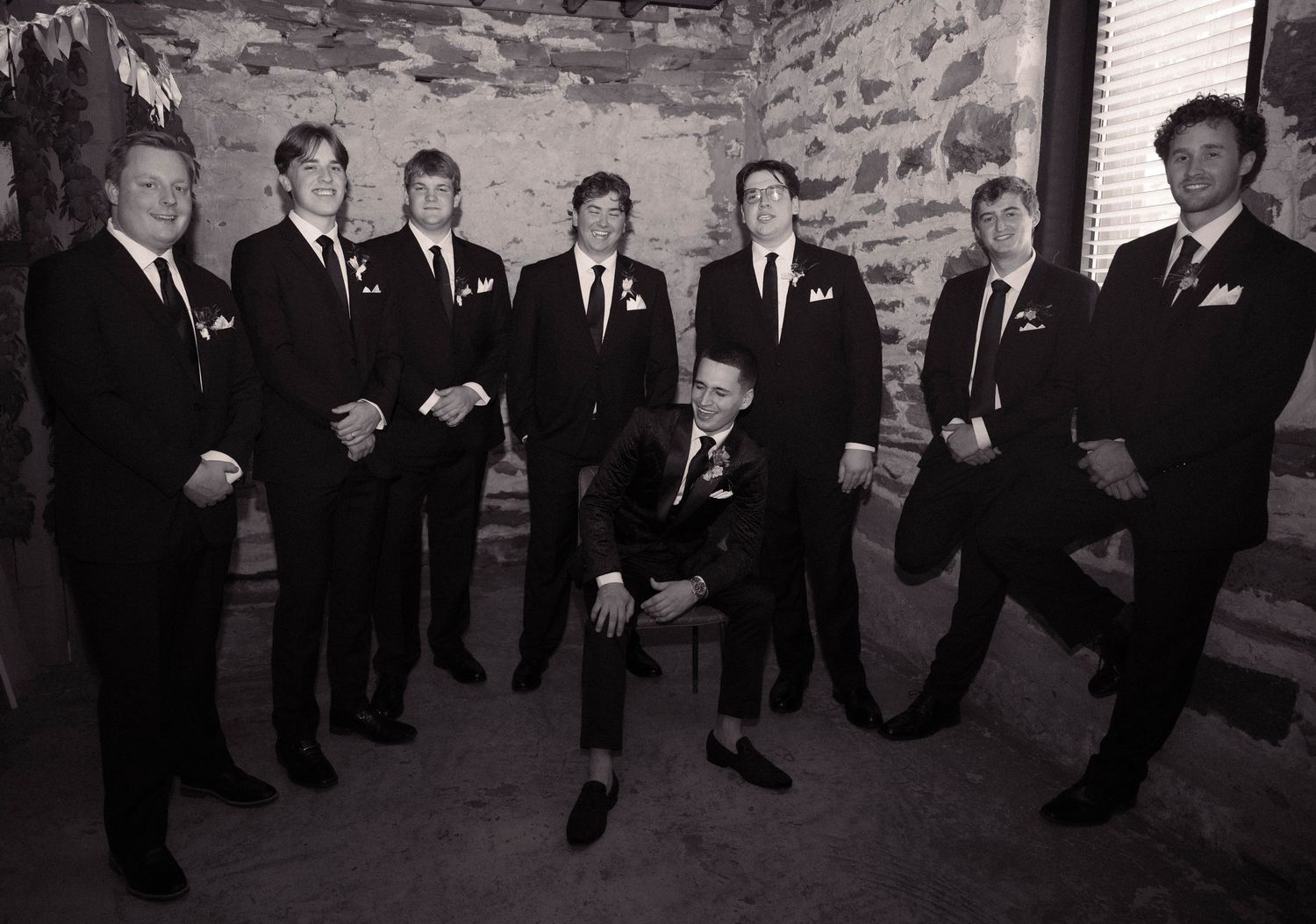 Black and white formal portrait of groom and groomsmen in suits against stone wall backdrop.