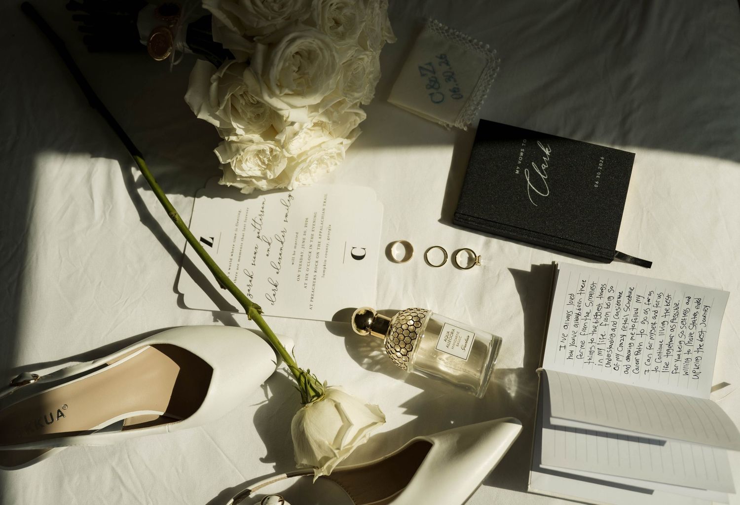 Flat lay of wedding details including rings, perfume, white roses, and vows on white surface.