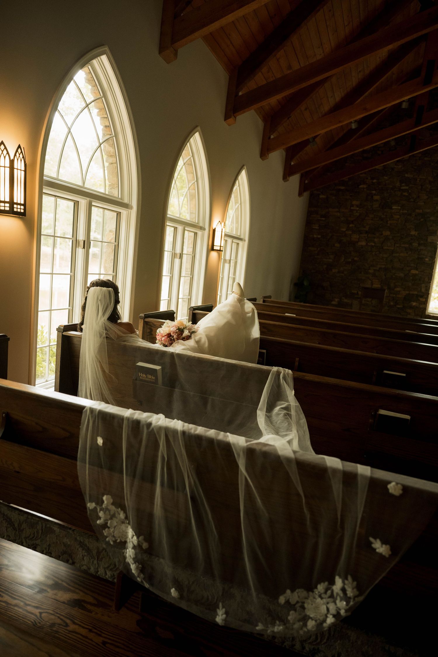 Bride sitting in church pew with long floral veil draped over wooden bench near arched windows.