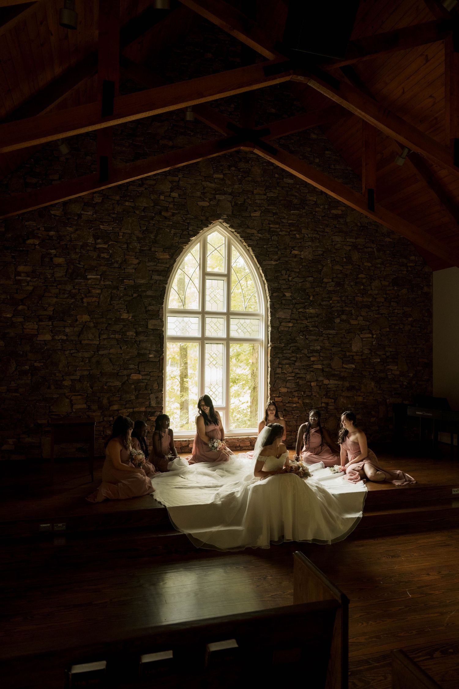 Bride and bridesmaids seated in circle on chapel floor bathed in light from gothic window.