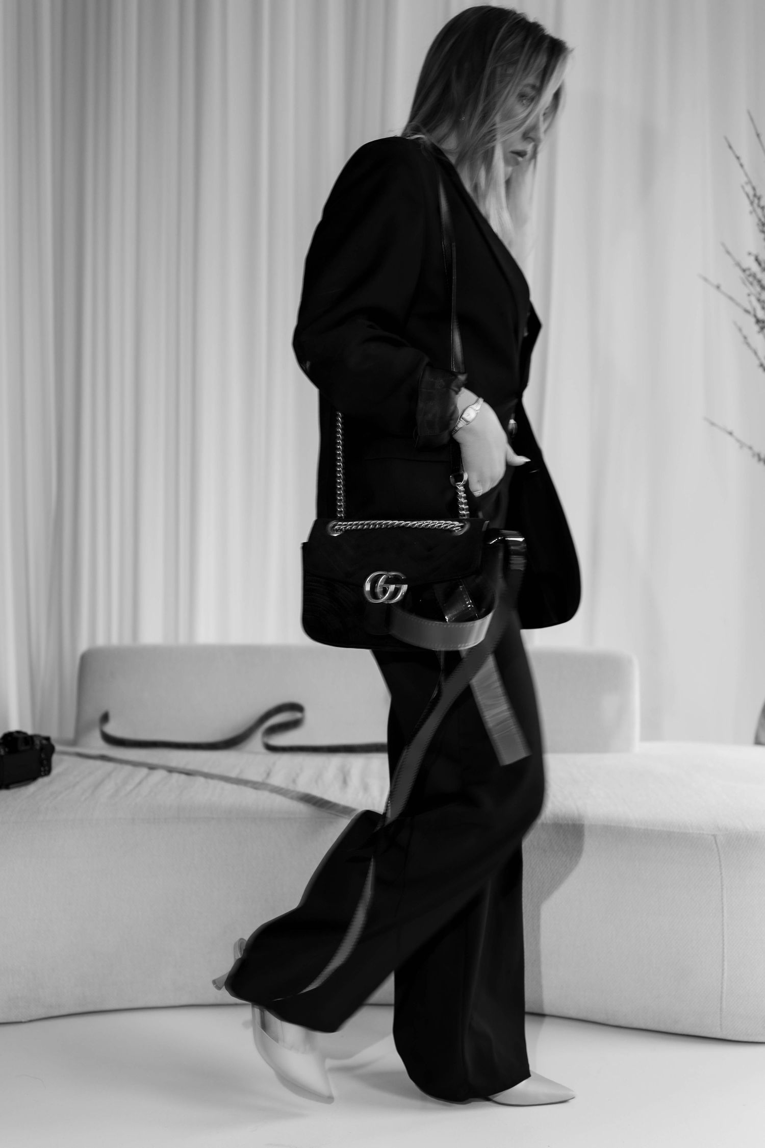 Black and white image of a woman in a black blazer carrying a Gucci chain shoulder bag with wide-leg pants.