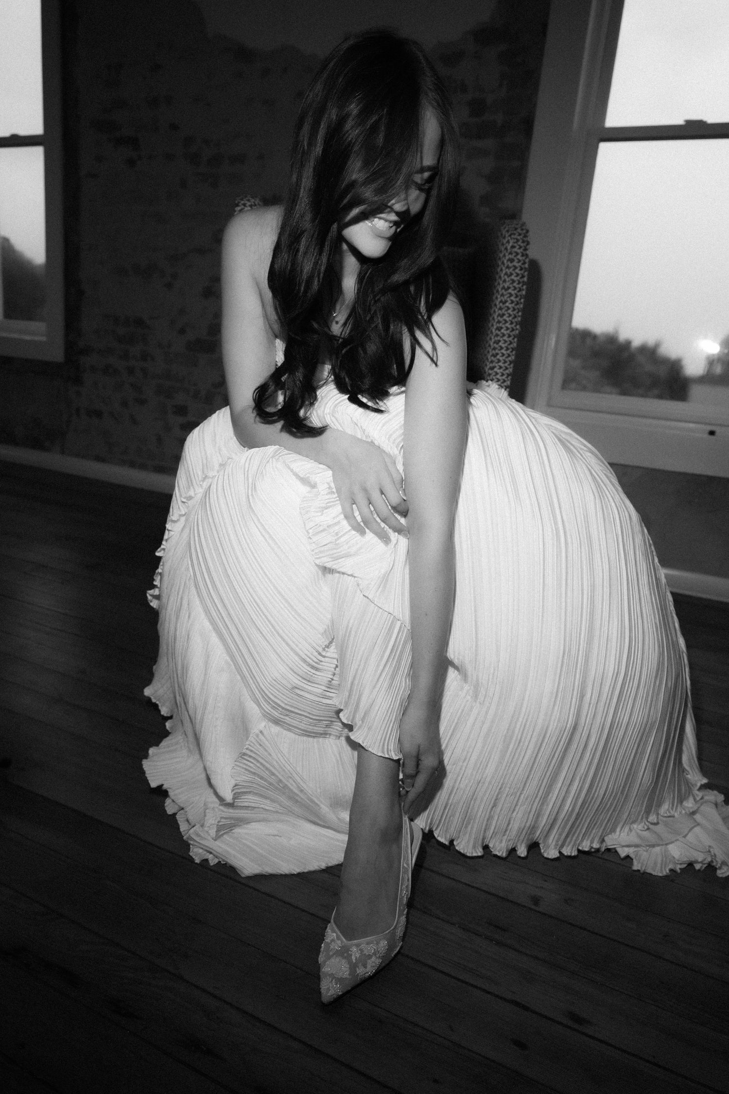 Black and white photo of a bride in a pleated gown adjusting her heel while seated indoors.