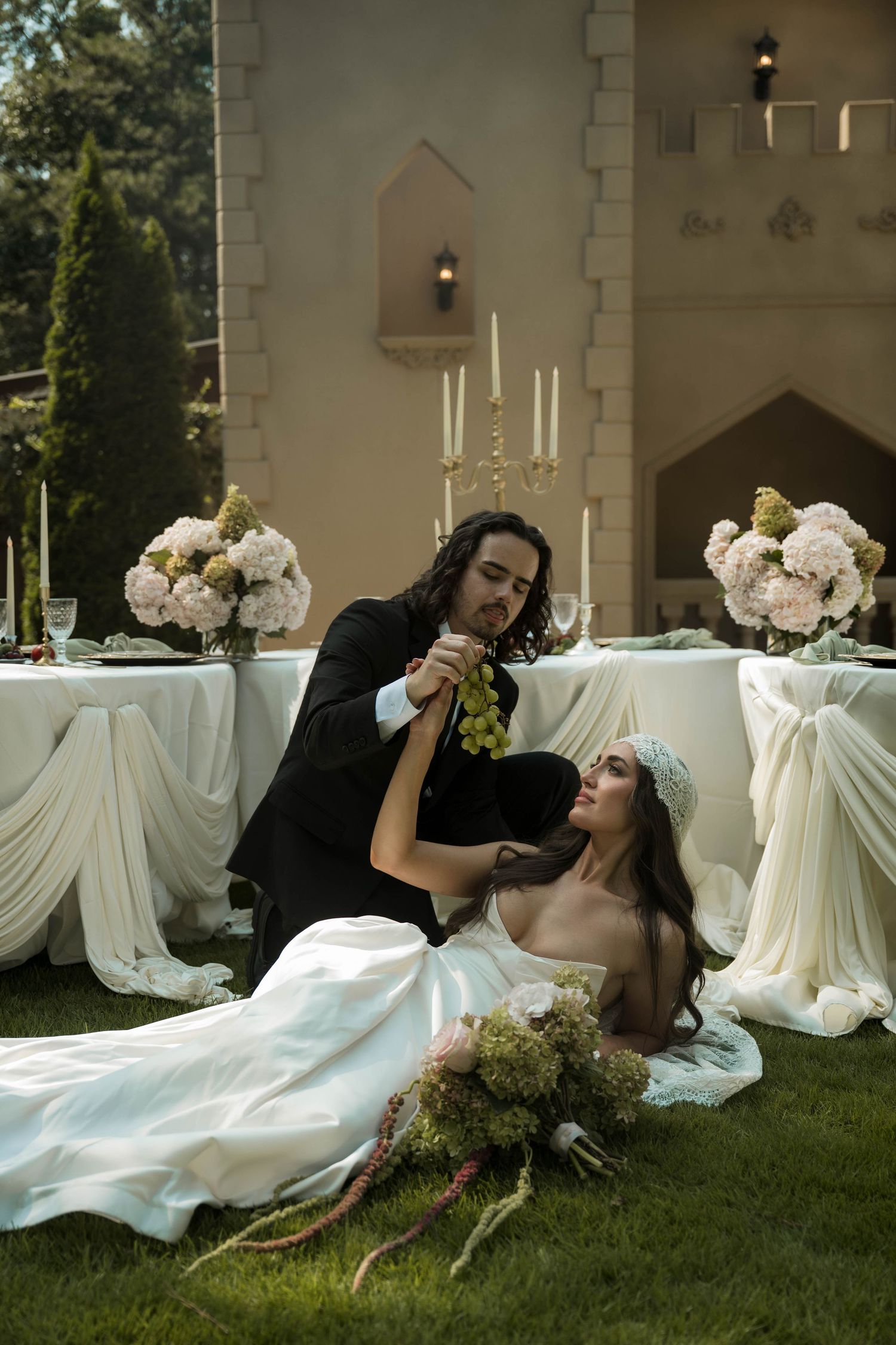 Romantic wedding couple outdoors, groom feeding bride grapes near elegant floral table setting with candles.