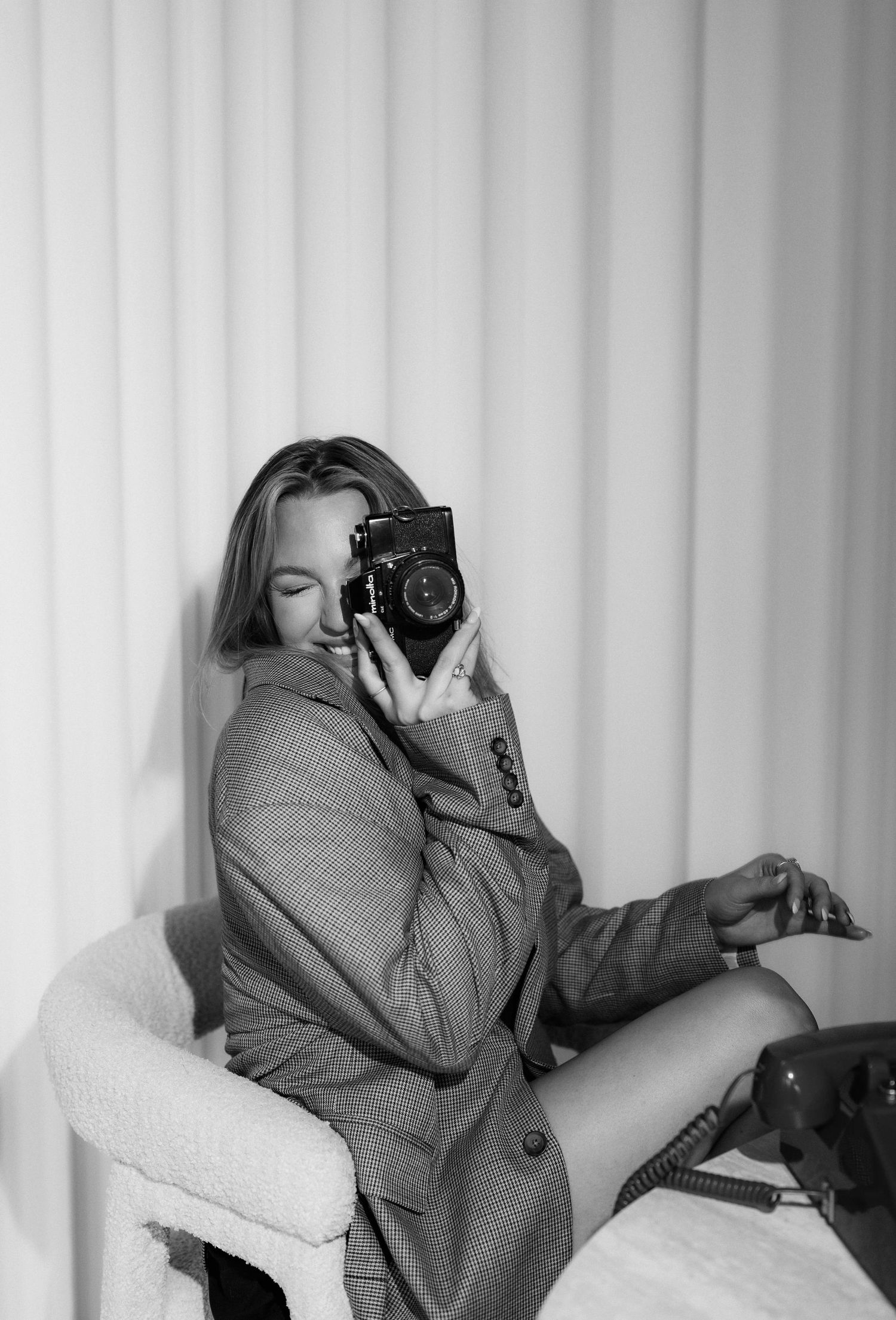 Black and white portrait of a woman in an oversized blazer holding a vintage film camera while seated.