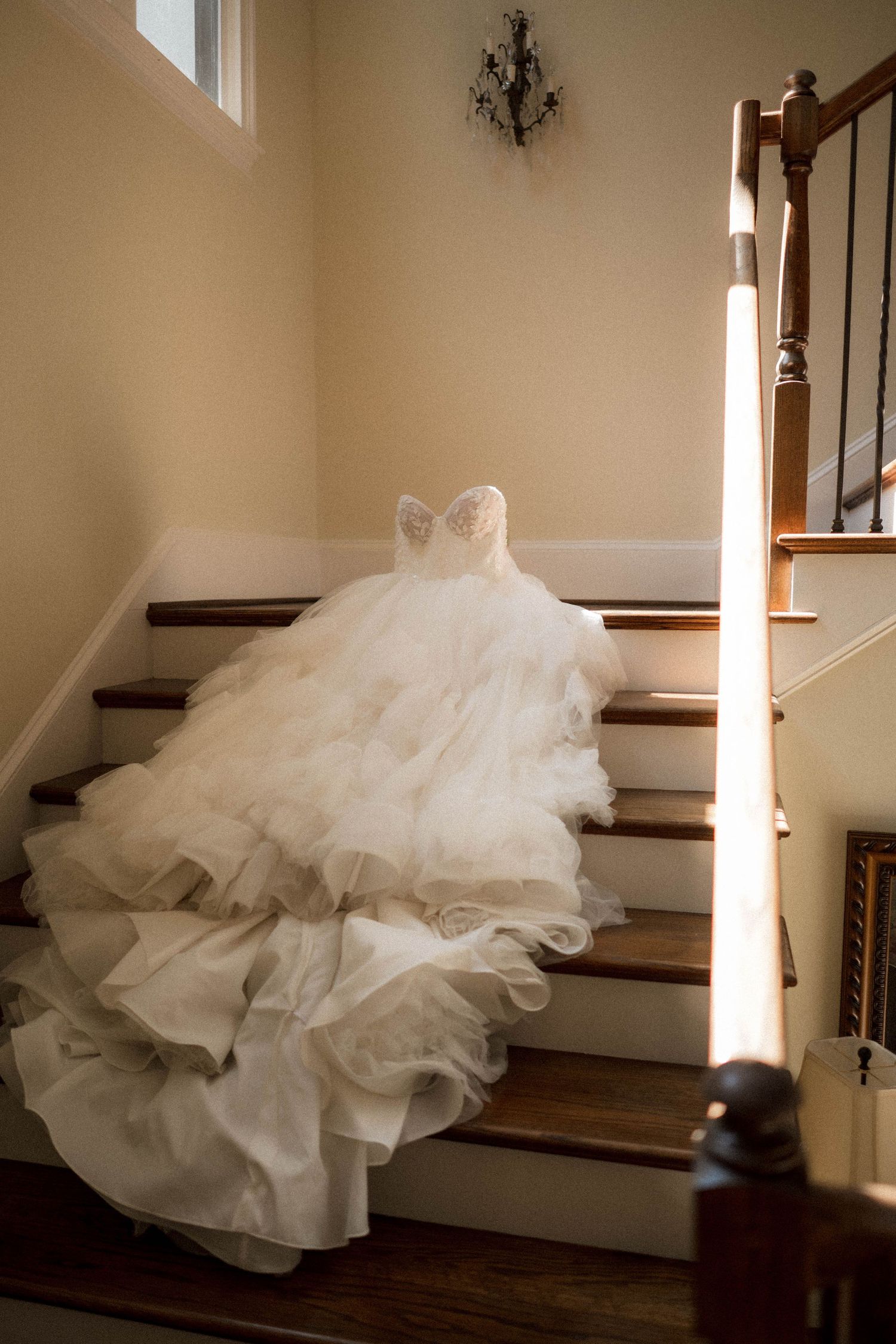 Elegant strapless ruffled wedding gown displayed on sunlit wooden staircase in warm interior.
