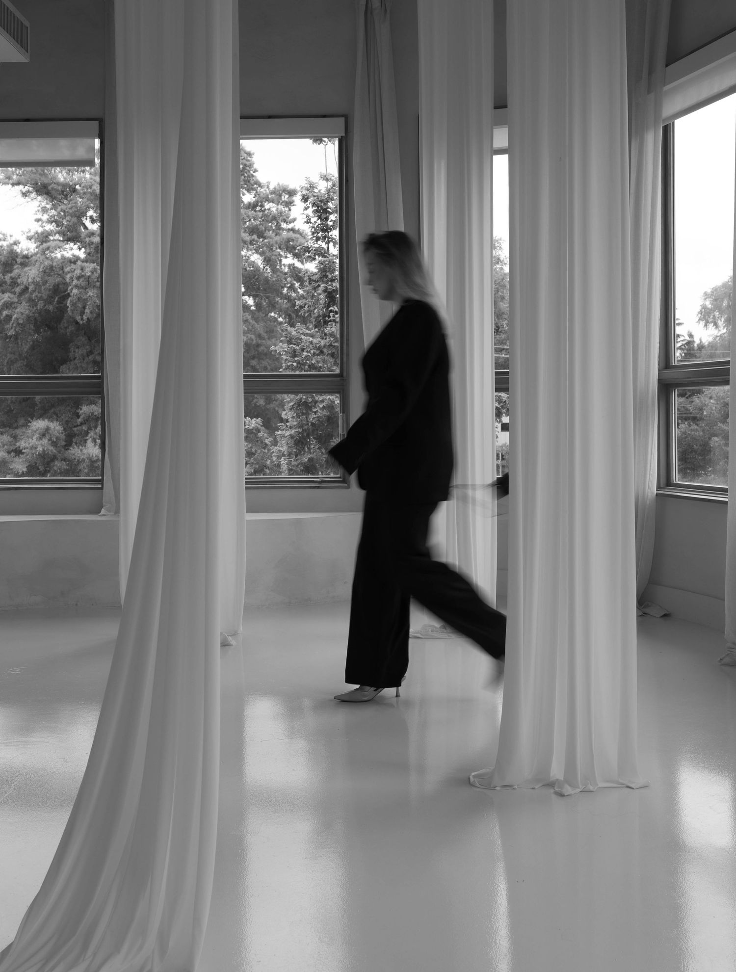 Blurred black and white photo of a woman in black walking between white draped curtains in a bright room.