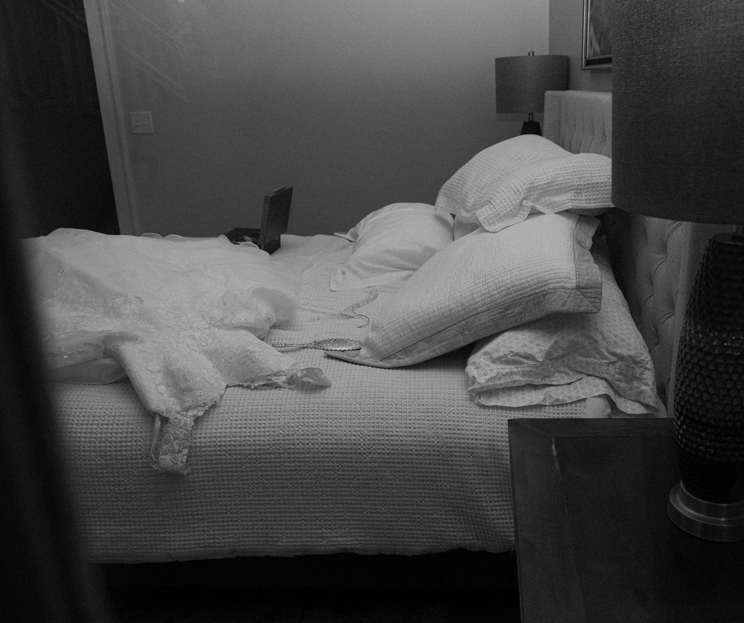 Black and white photo of unmade bed with rumpled white pillows and sheets in dimly lit bedroom.