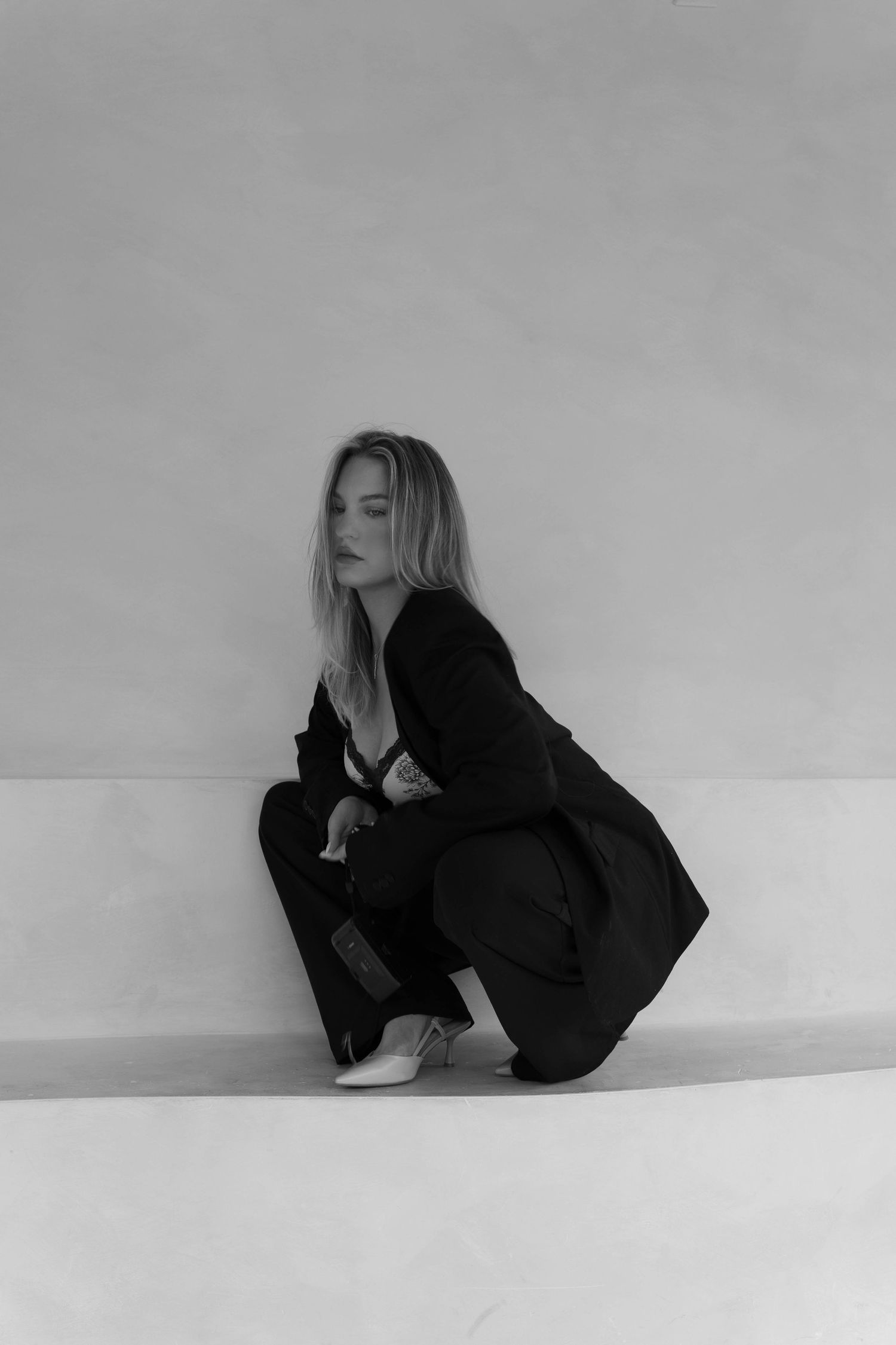 Black and white photo of a blonde woman in a black suit crouching against a light-colored wall.