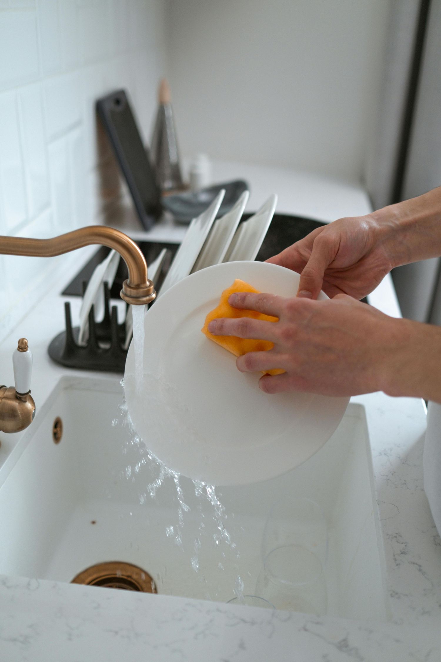 Hands washing a white plate in a kitchen sink with yellow sponge and brass faucet.