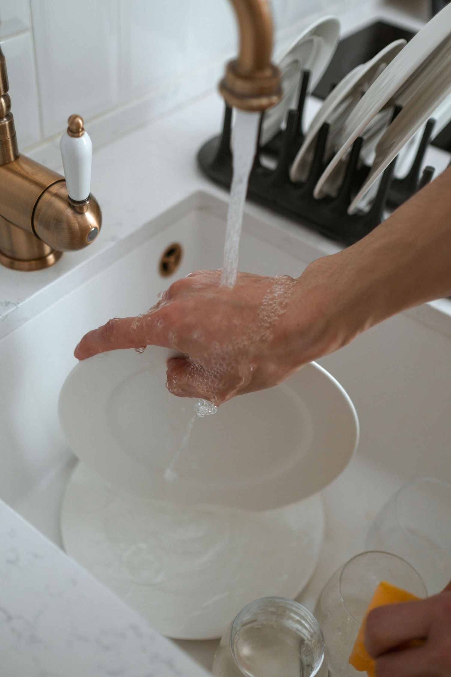 Hands washing dishes in a white ceramic sink with running water from a gold-toned faucet.
