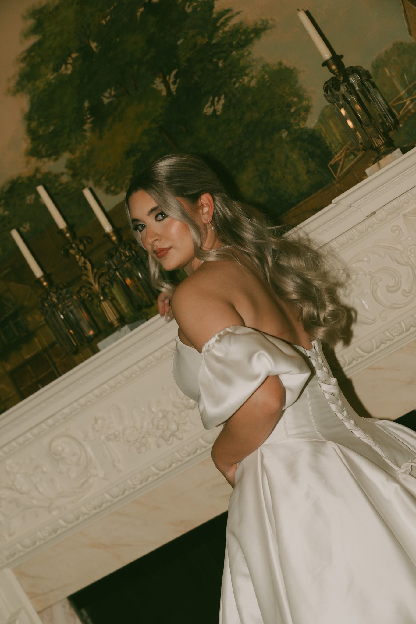 A person in a luxurious white satin wedding gown poses elegantly against an ornate interior backdrop.