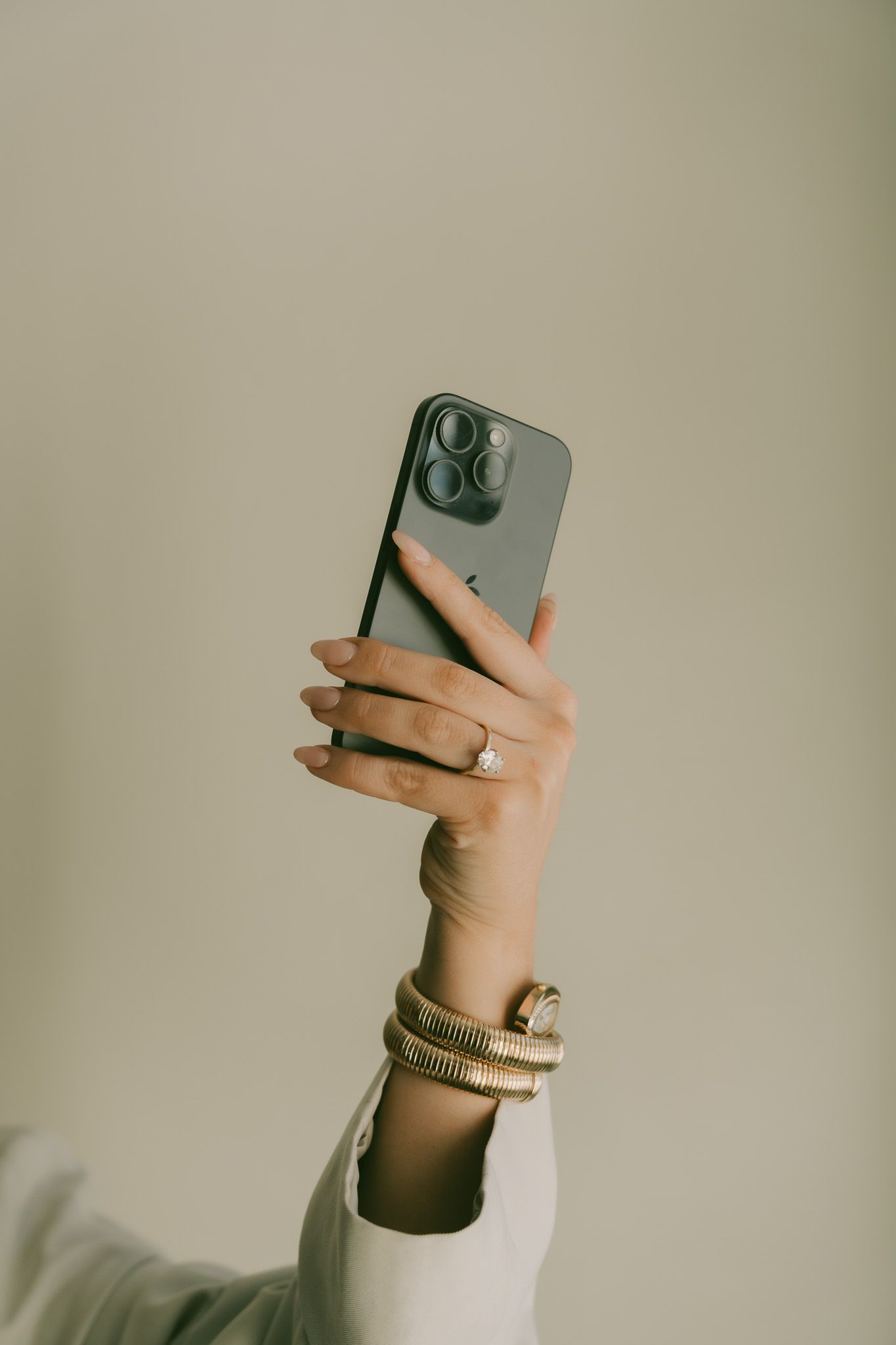 Hand holding an iPhone while wearing a cream sweater and gold bracelet against a neutral beige background.
