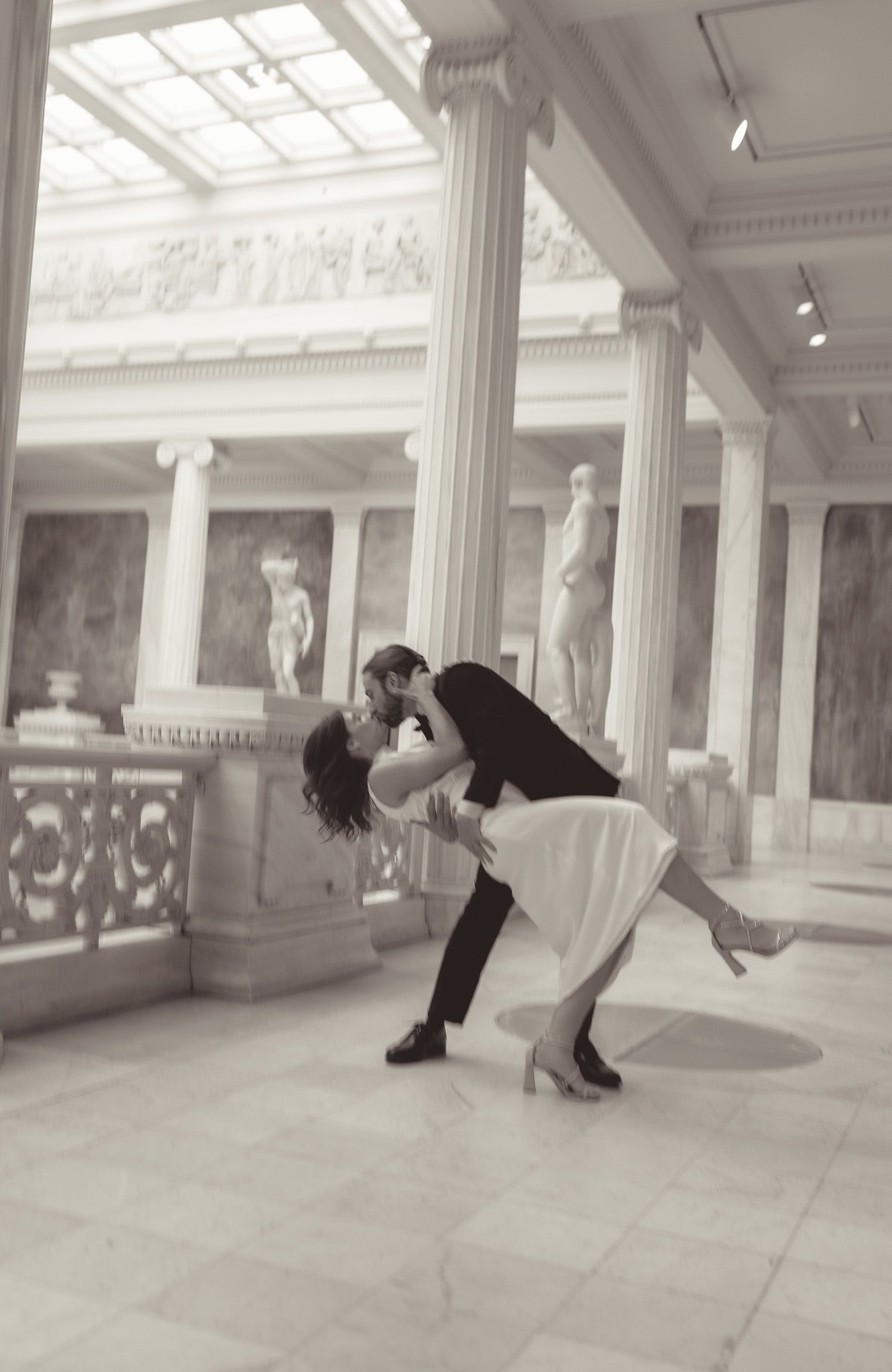 couple does a dip kiss across a grand hall with ornate white columns and skylights in carnegie museum of art in pittsburgh.