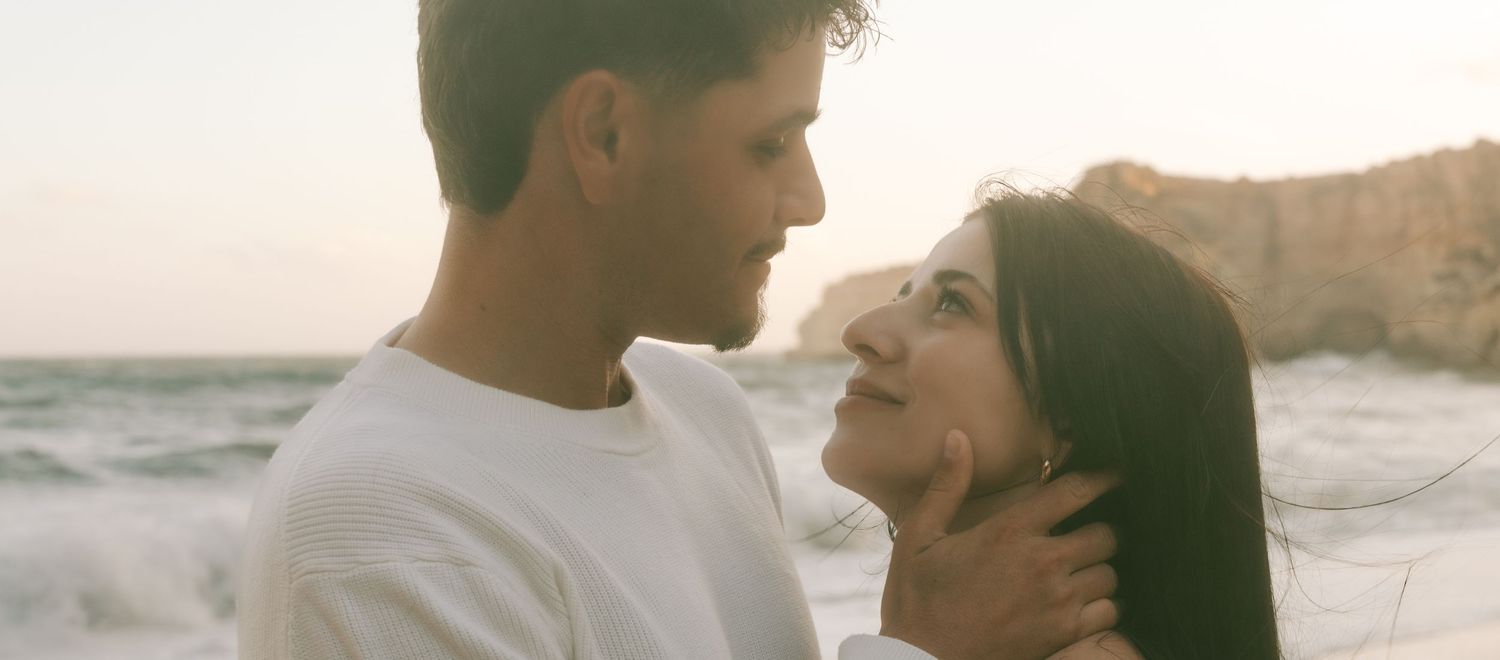 Romantic couple sharing a tender moment on a beach at sunset, man gently holding woman's face near rocky cliffs.