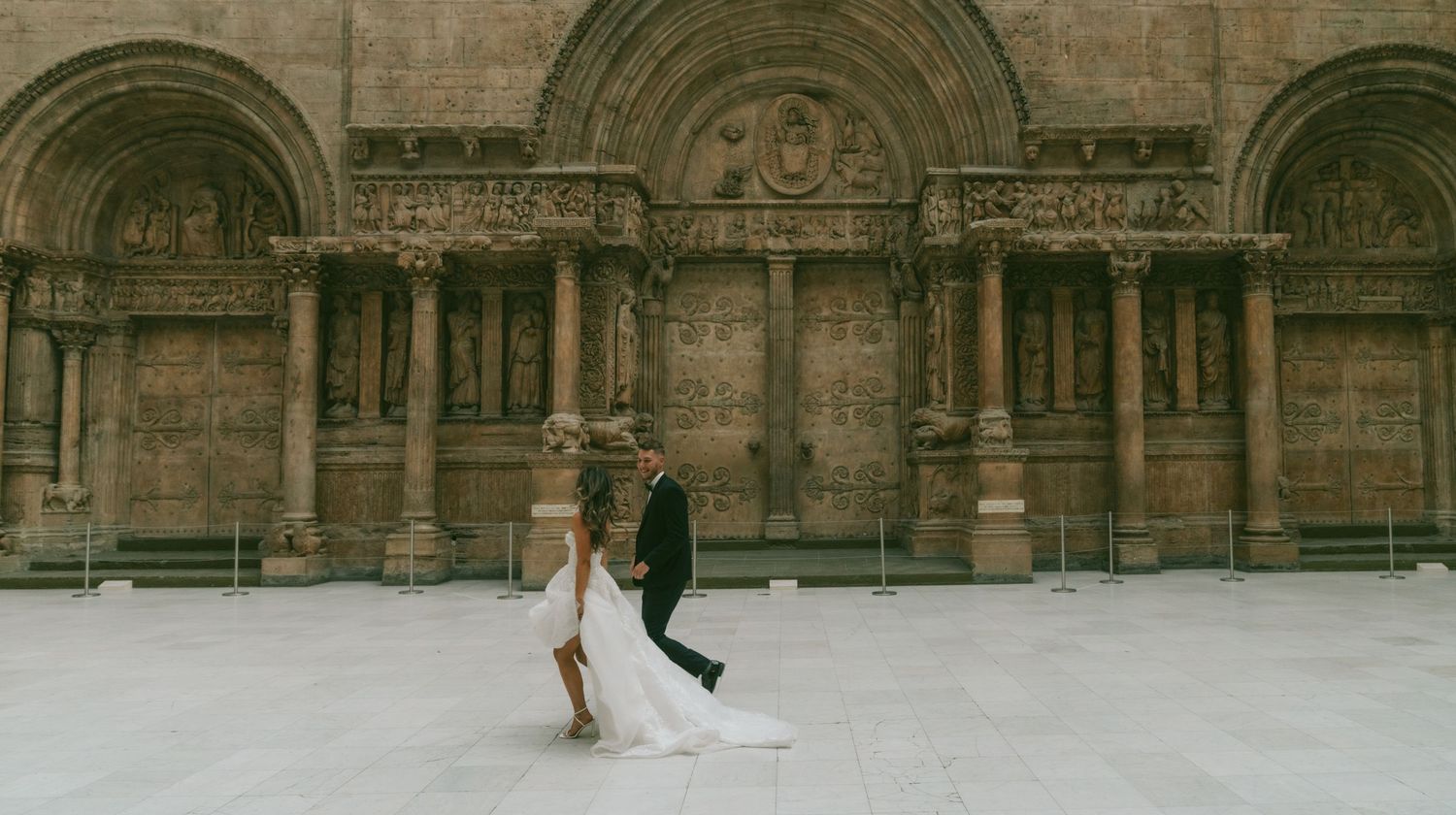 A bride and groom walk hand in hand along a historic stone building facade with ornate architectural details.