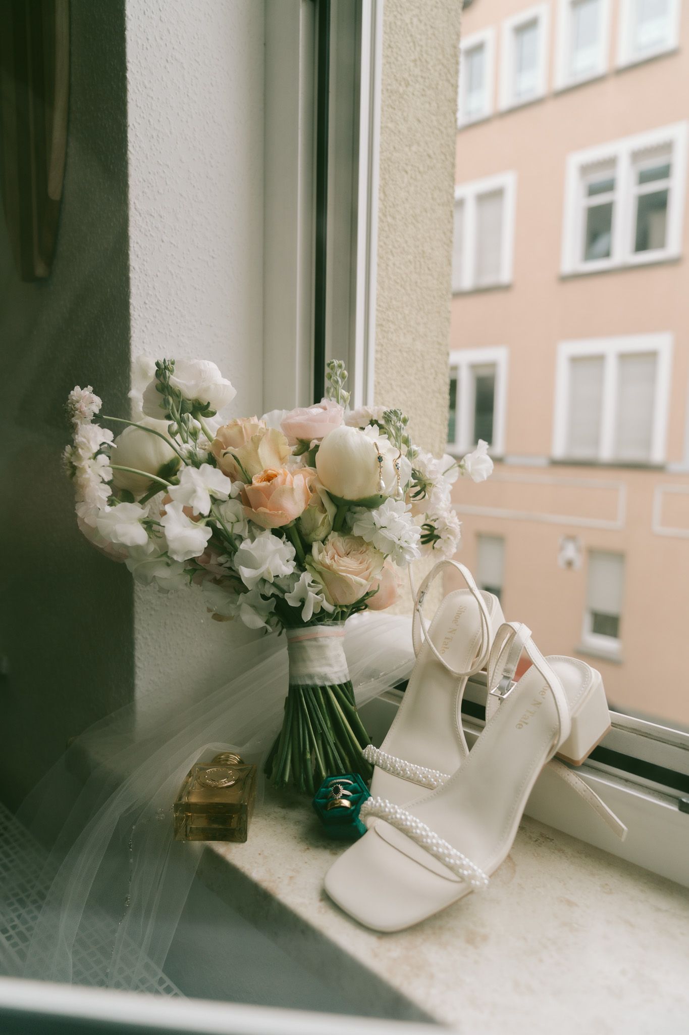 A delicate bridal bouquet of white and peach roses sits on a windowsill next to elegant white wedding shoes in Germany.
