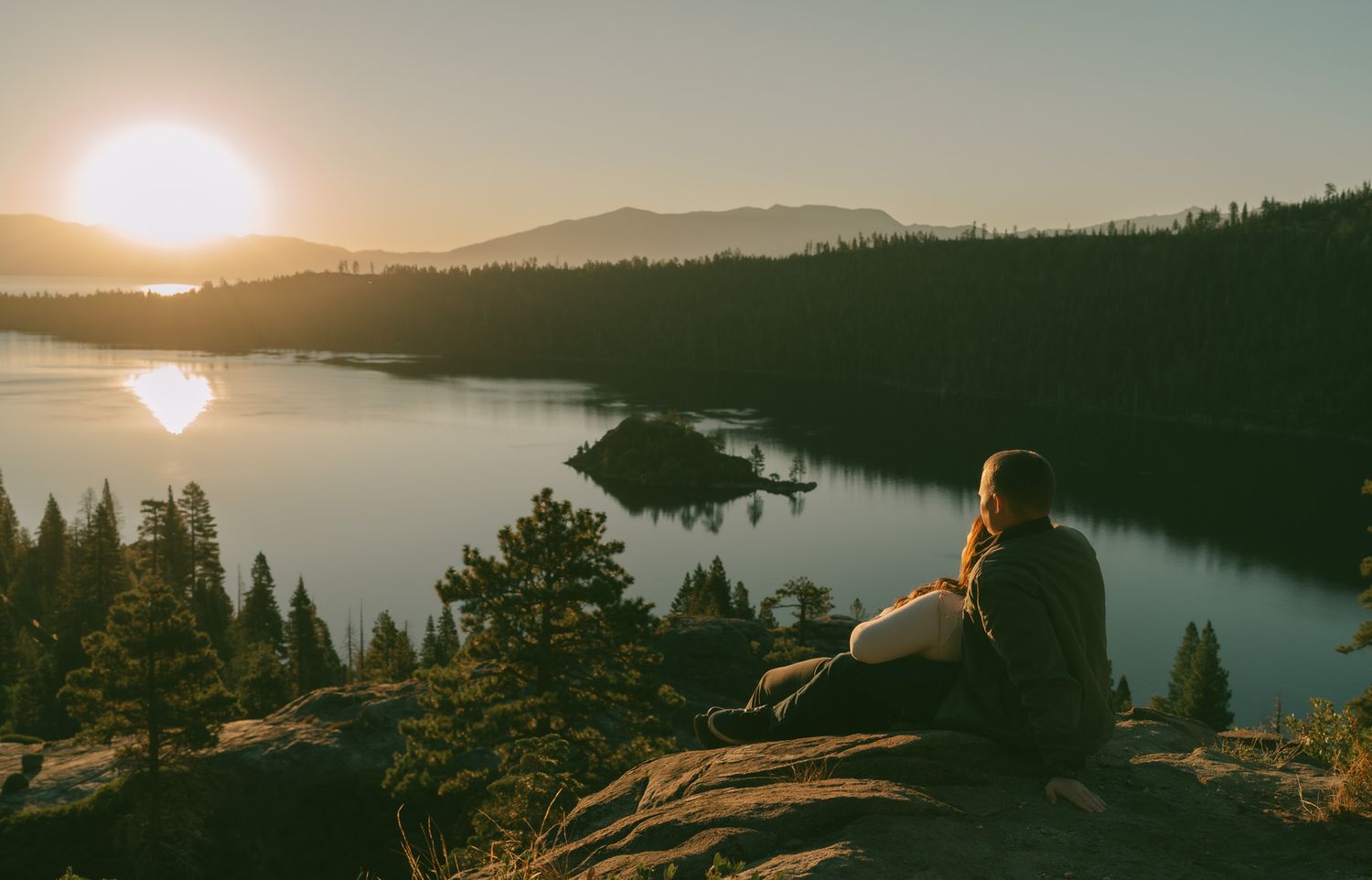 Panoramic sunset view over a lake surrounded by forested mountains.