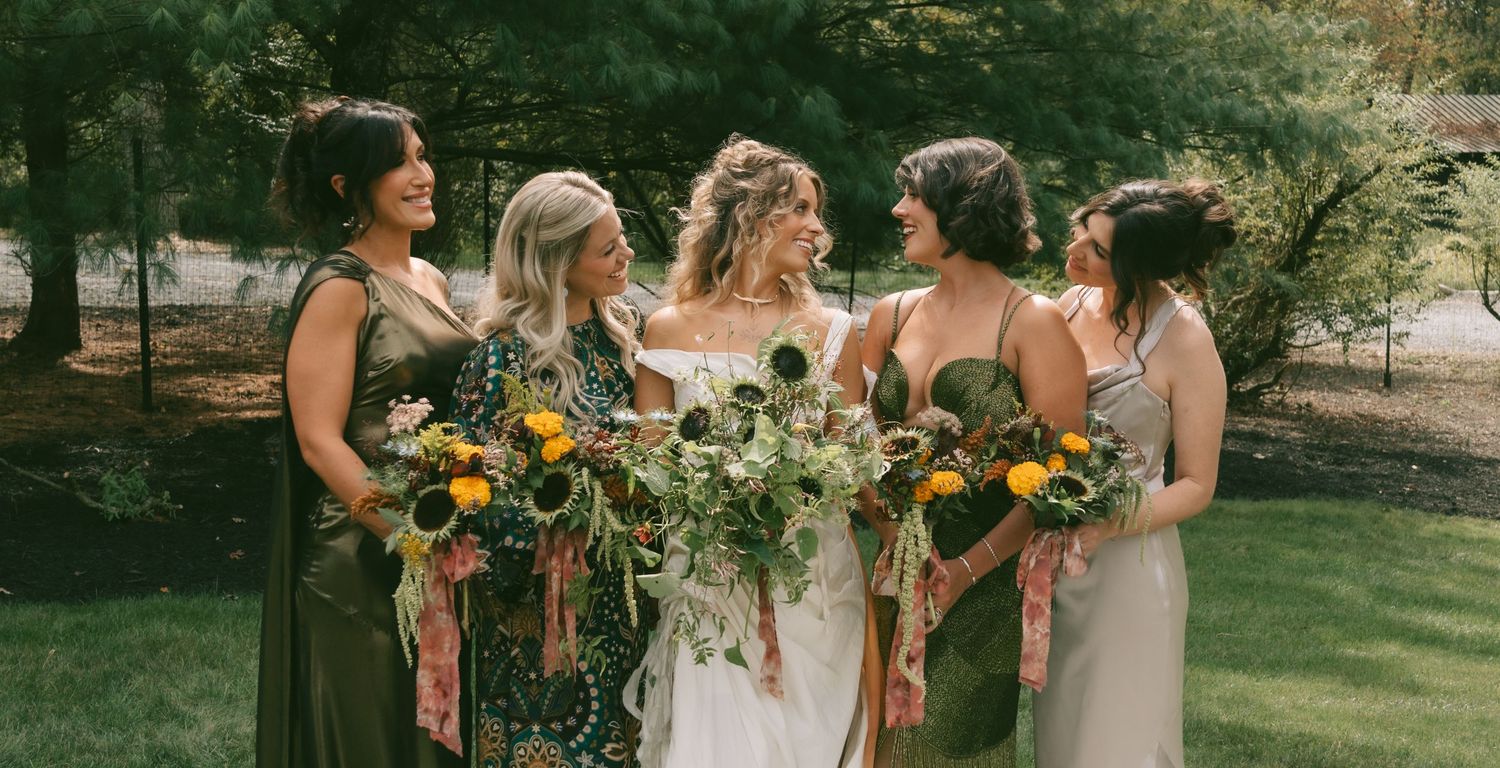Bridesmaids in champagne dresses holding vibrant sunflower and wildflower bouquets pose together outdoors.