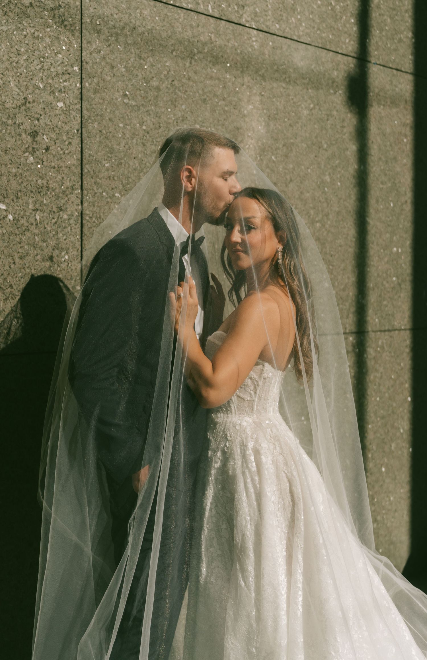 A romantic wedding couple shares an intimate moment against a concrete wall with dramatic shadows and flowing veil.