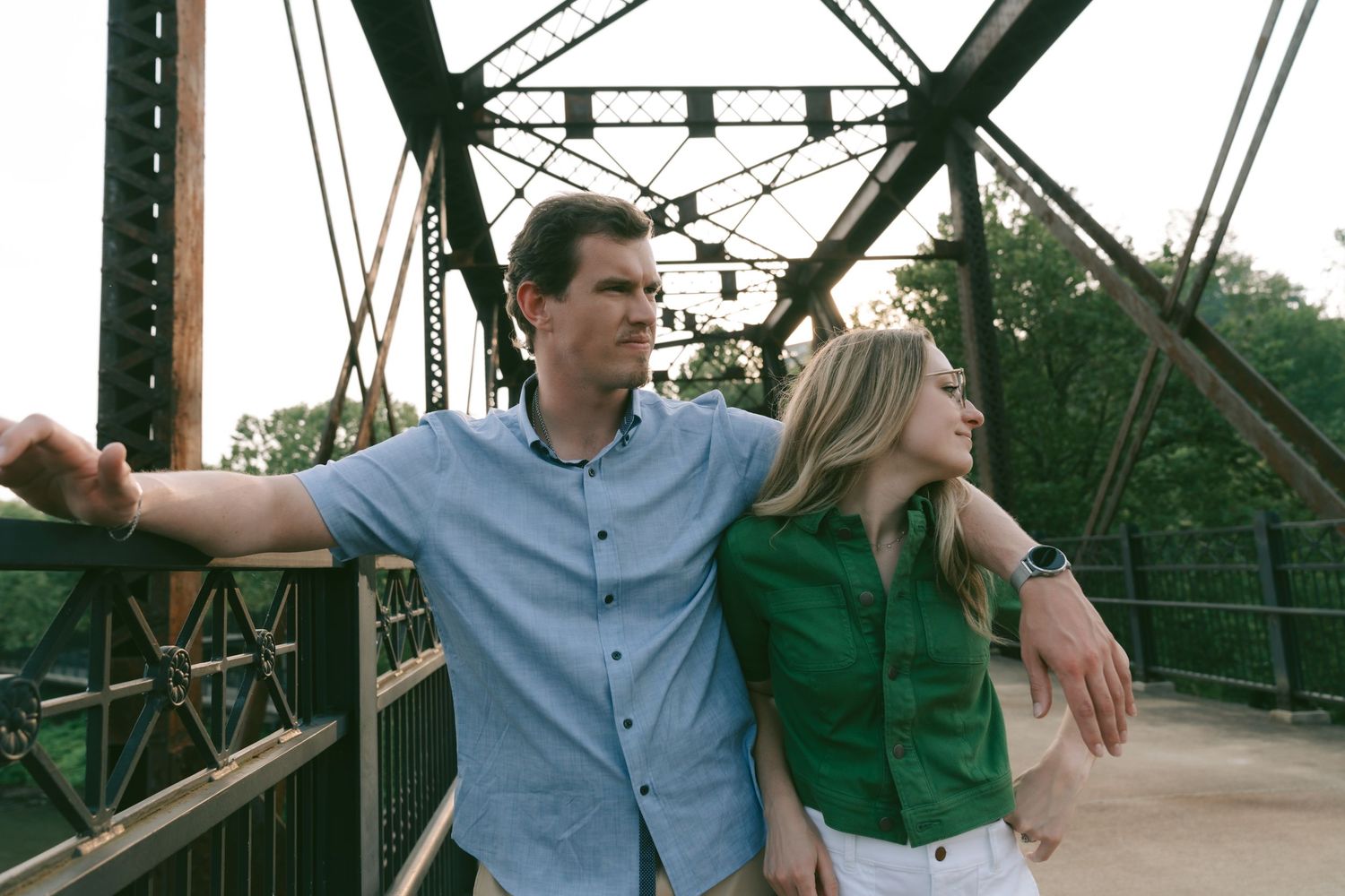 A couple in casual attire walks and poses together on a rustic metal bridge structure in a natural setting.