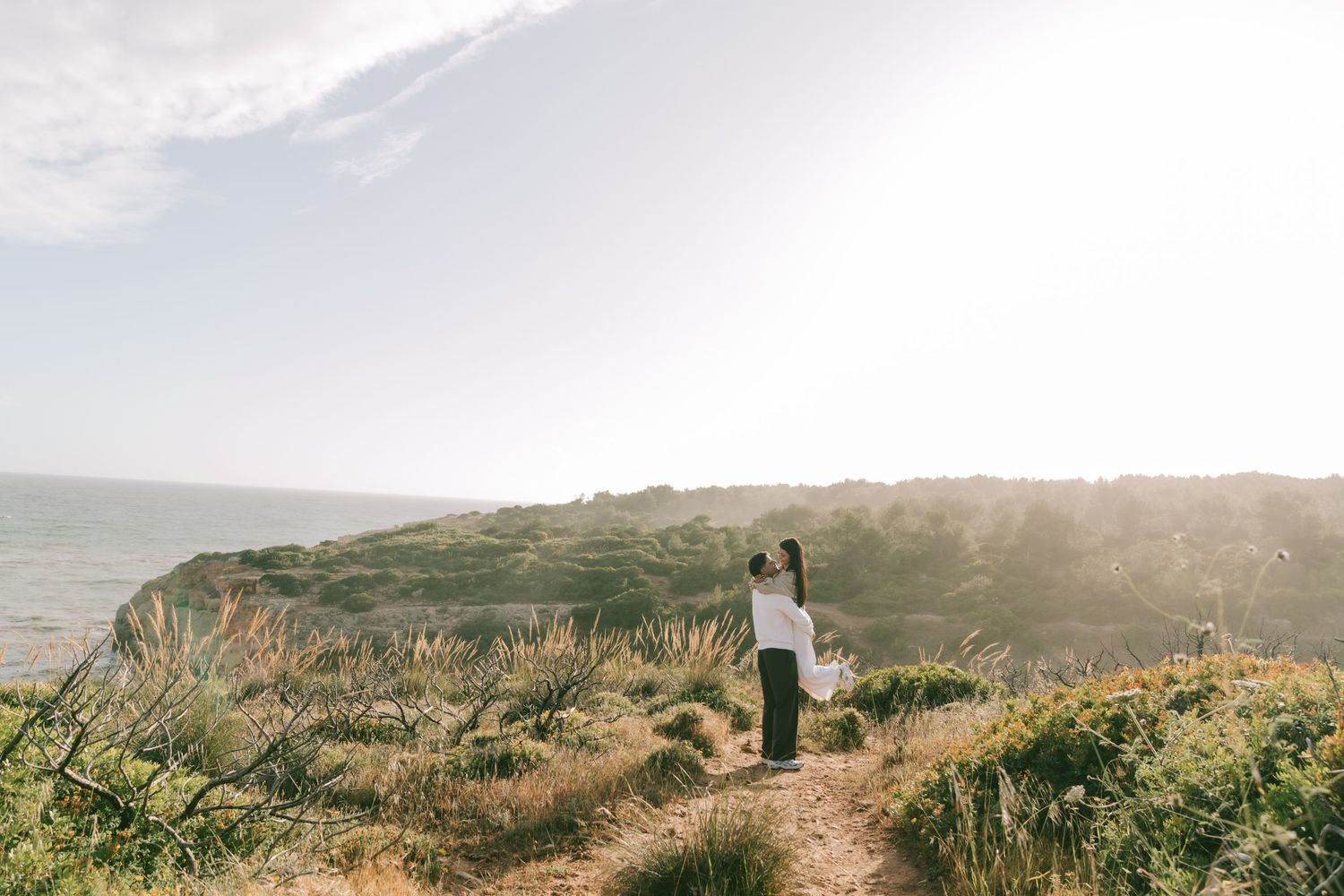 A woman stands on a scenic coastal trail overlooking lush green hills and the ocean in golden sunlight.