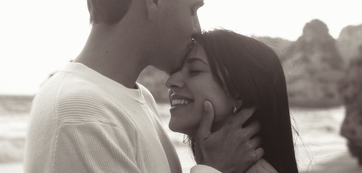 Black and white photo of a romantic couple sharing a tender moment, smiling and embracing on a beach.