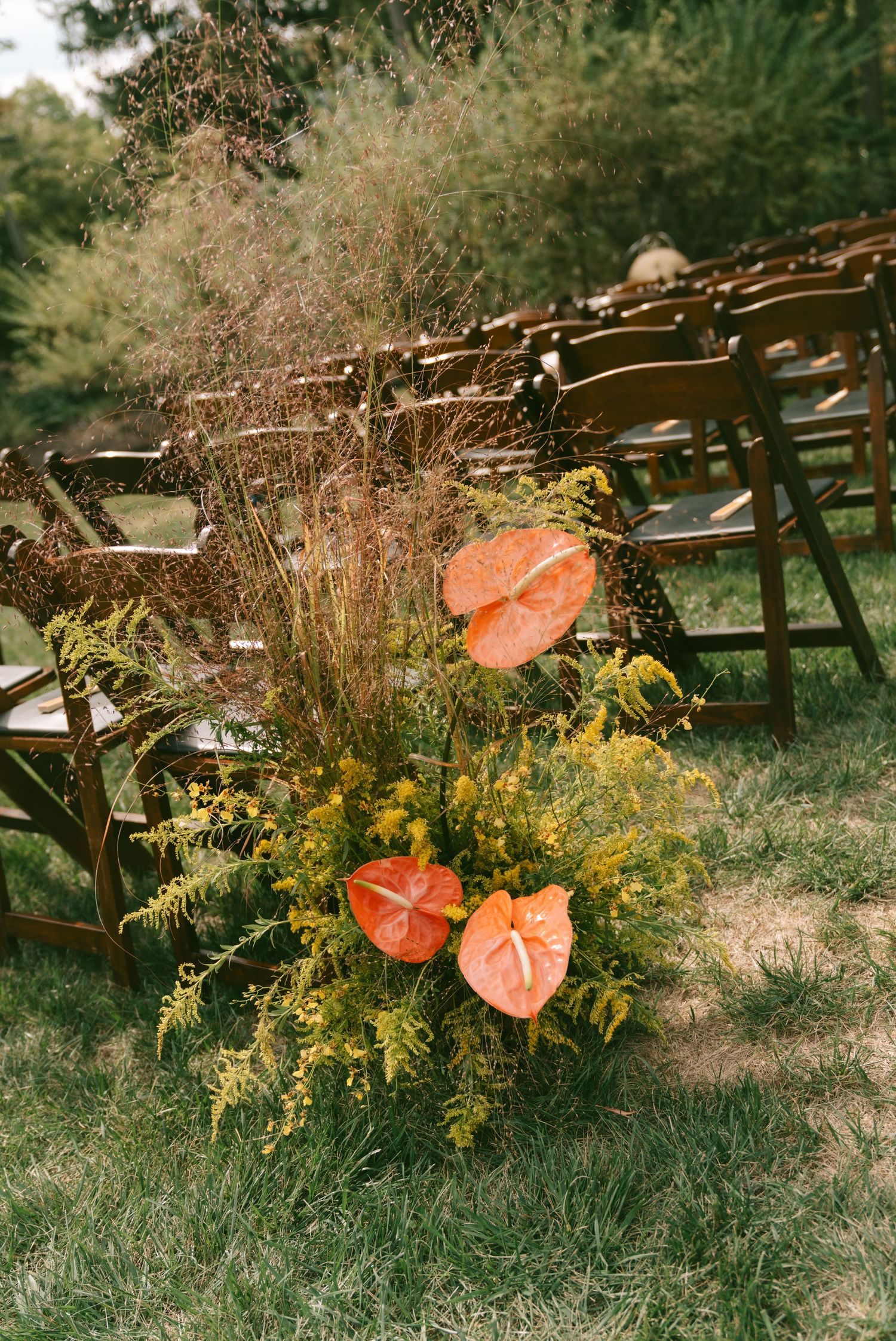Coral-colored floral arrangements with yellow wildflowers decorate wooden chairs at an outdoor wedding ceremony.