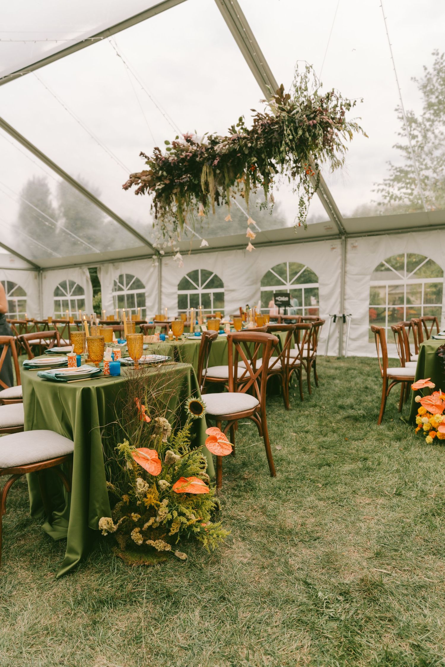 Wedding reception tent setup with wooden chairs and orange floral arrangements under clear ceiling panels.