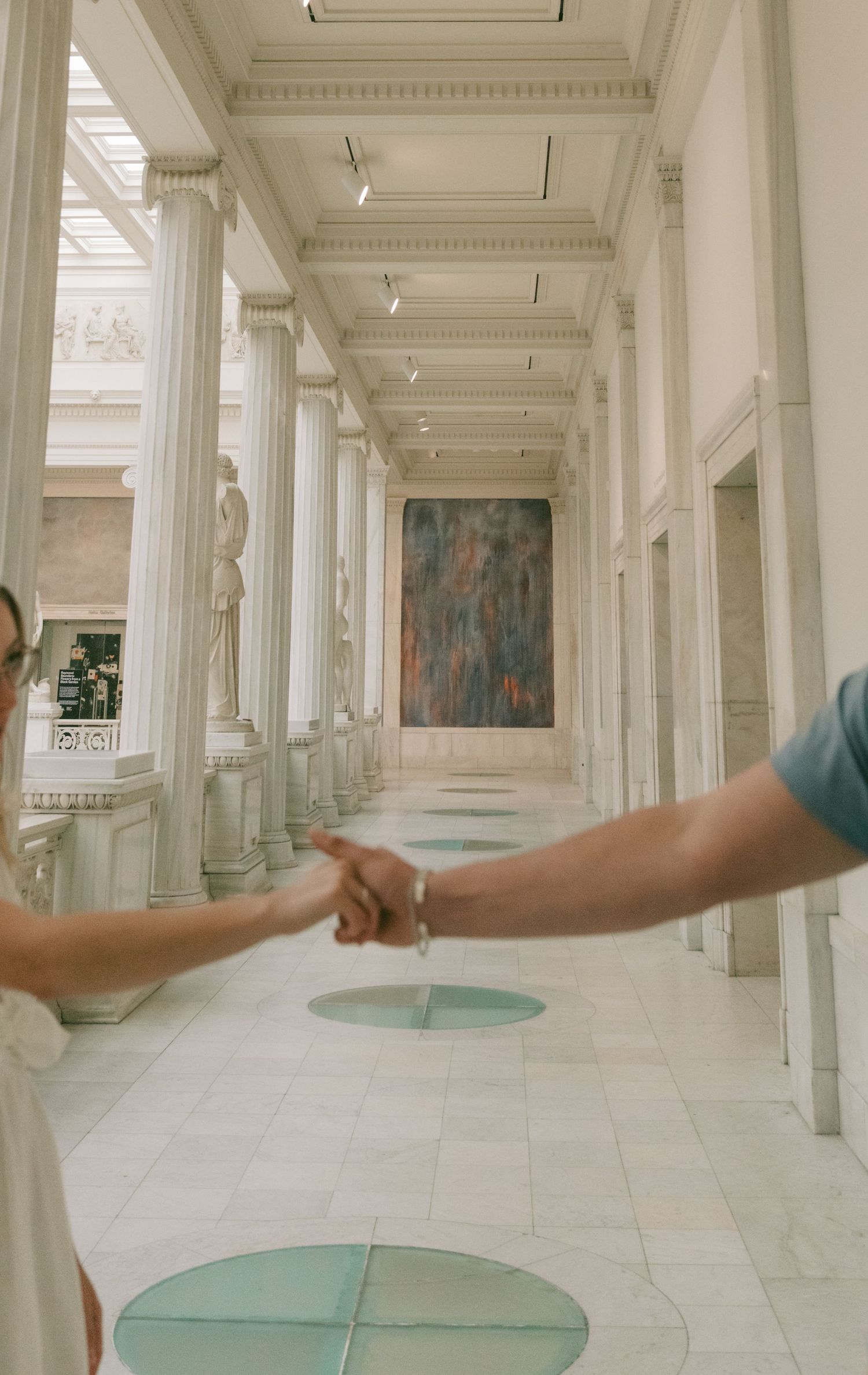 Two people hold hands in a grand white hallway with classical columns and circular floor patterns.